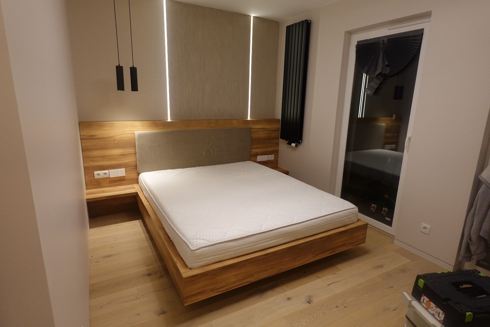 Drewniane łóżko z tapicerowanym zagłówkiem, oświetleniem LED i szafkami nocnymi wbudowanymi w ramę. Widoczny grzejnik i okno z roletą. Całość w nowoczesnym stylu.