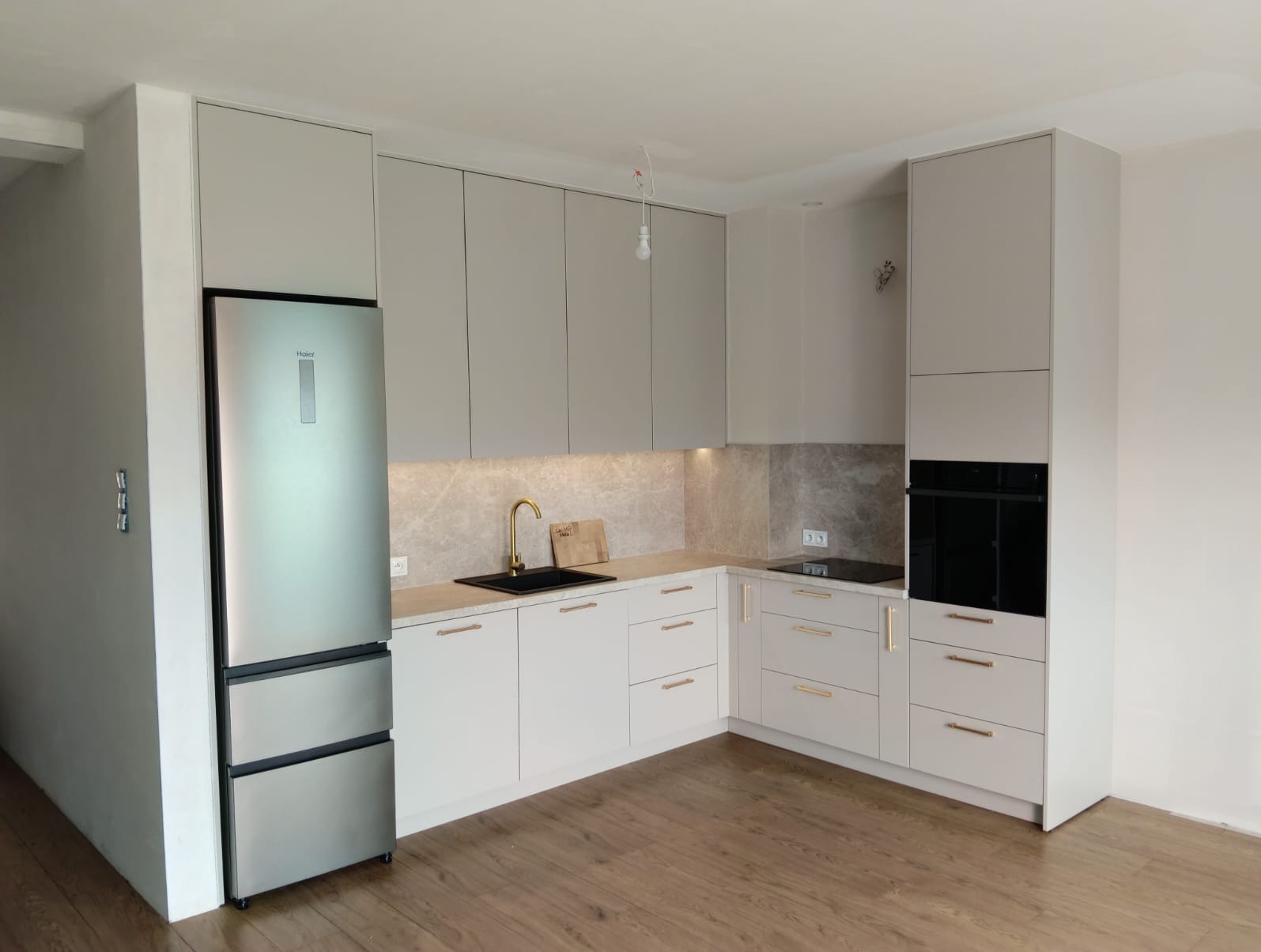 Jasna kuchnia z szarymi szafkami i złotą armaturą. Nowoczesny projekt z lodówką Haier, drewnianą podłogą i subtelnym oświetleniem podszafkowym.