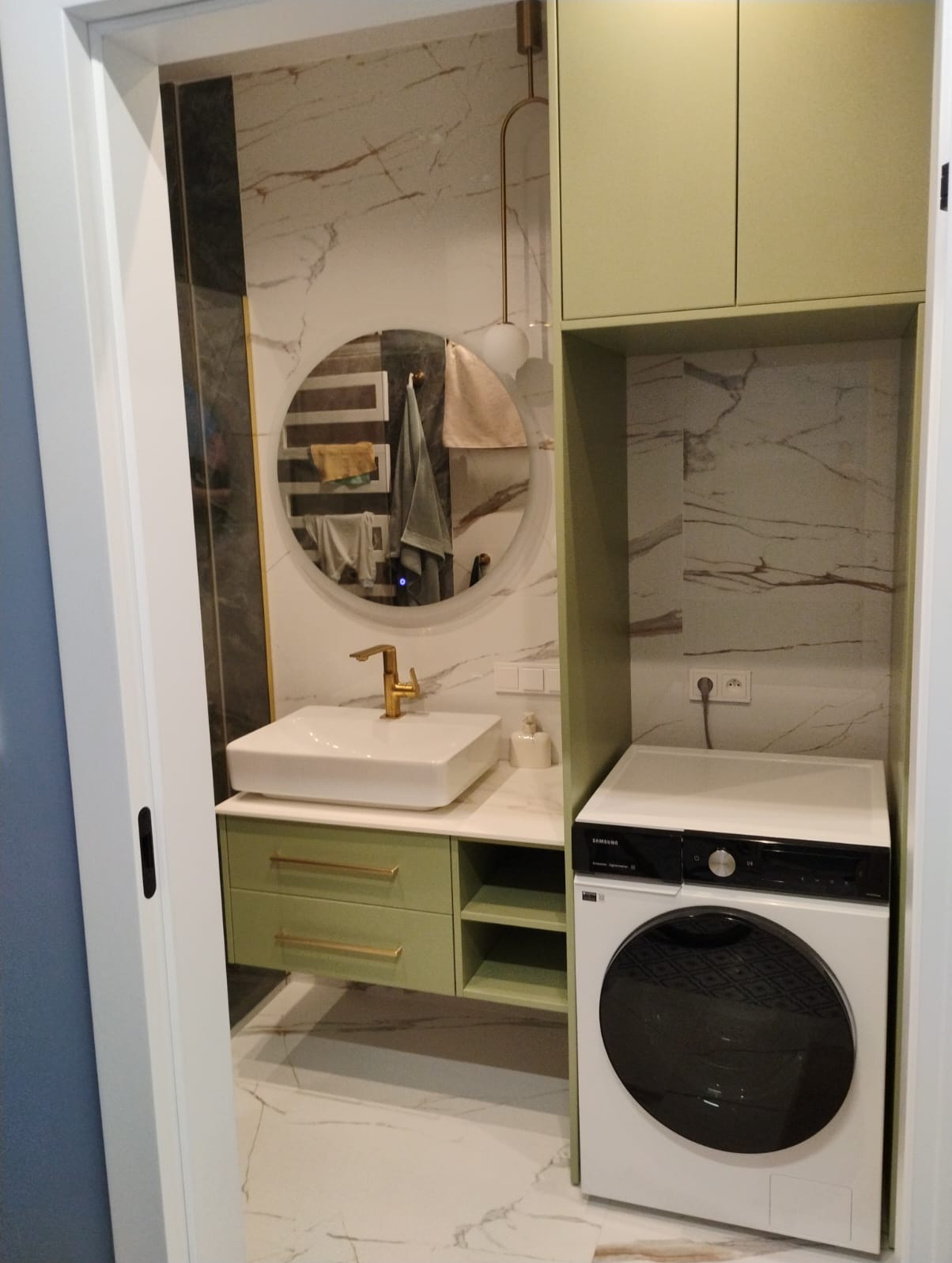 Nowoczesna łazienka z umywalką na szafce w odcieniach zieleni, okrągłym lustrem i pralką. Marmurowe płytki na ścianach i podłodze dodają elegancji.
