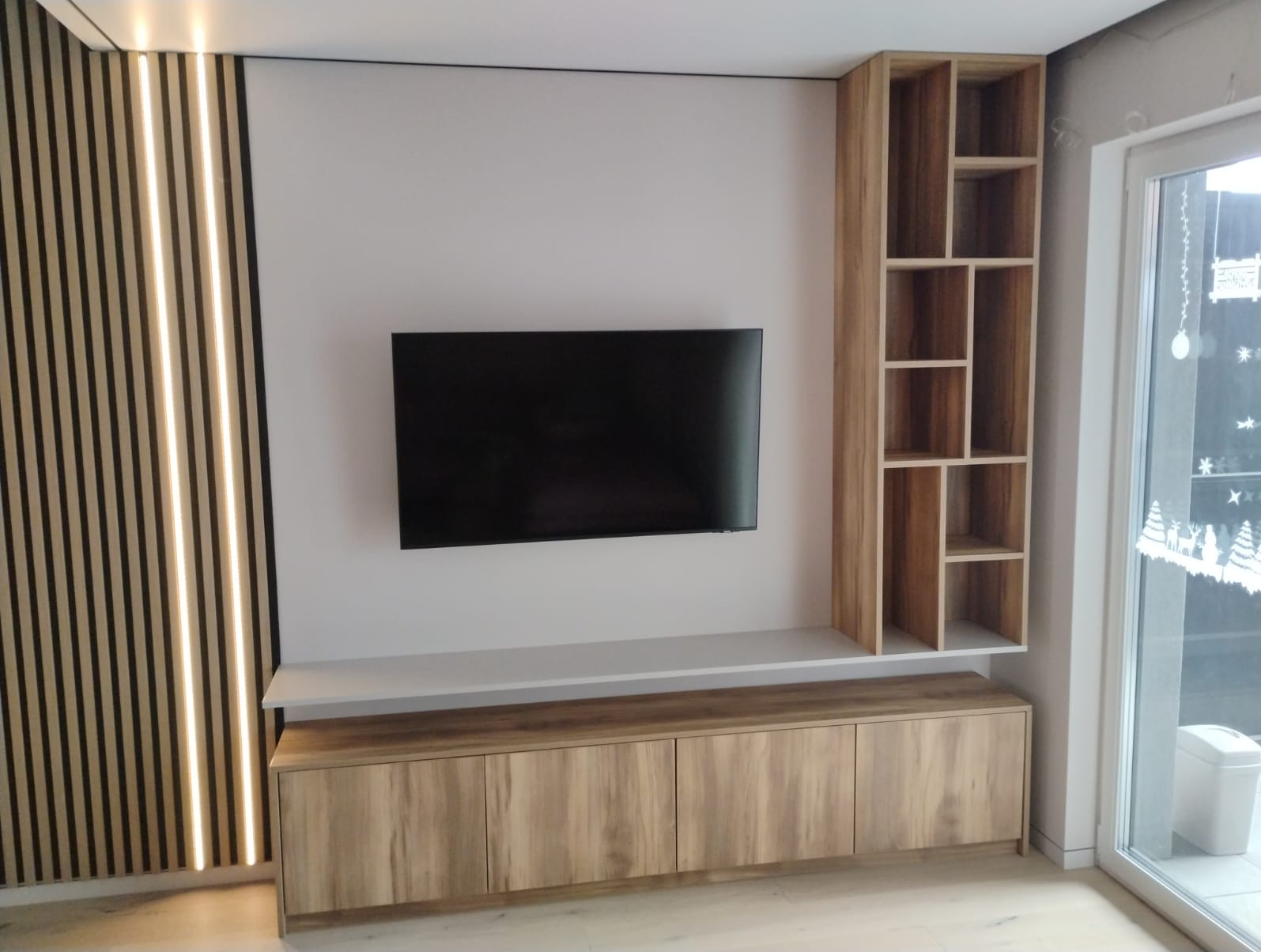 Nowoczesna zabudowa TV z lamelami LED, szafką RTV i regałem z jasnego drewna, tworząca spójną aranżację ściany w salonie. Minimalistyczny design.