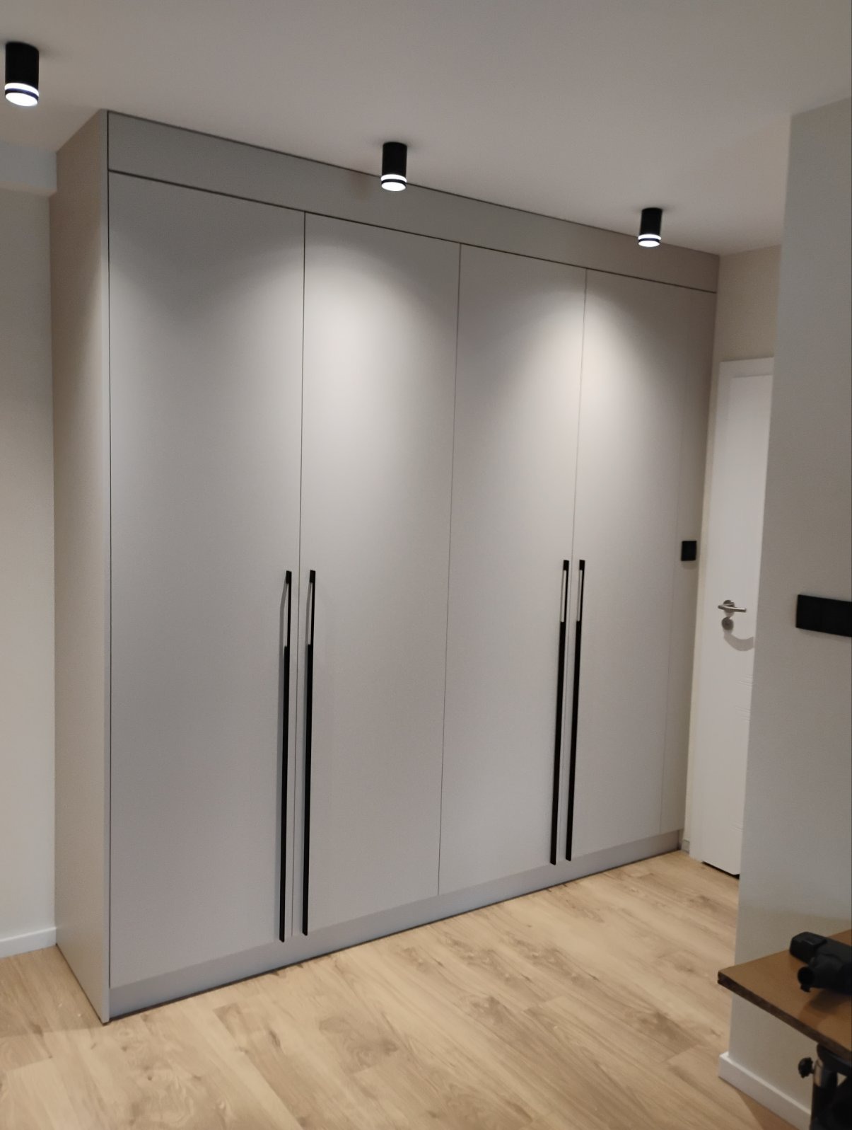Szara, nowoczesna szafa wnękowa z czarnymi, pionowymi uchwytami, oświetlona punktowo. Widoczna drewniana podłoga i fragment białej ściany z drzwiami.