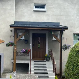 Zadaszone wejście do domu z drewnianą konstrukcją, czarną blachodachówką, wiszącymi kwiatami i schodami z antypoślizgowymi nakładkami.