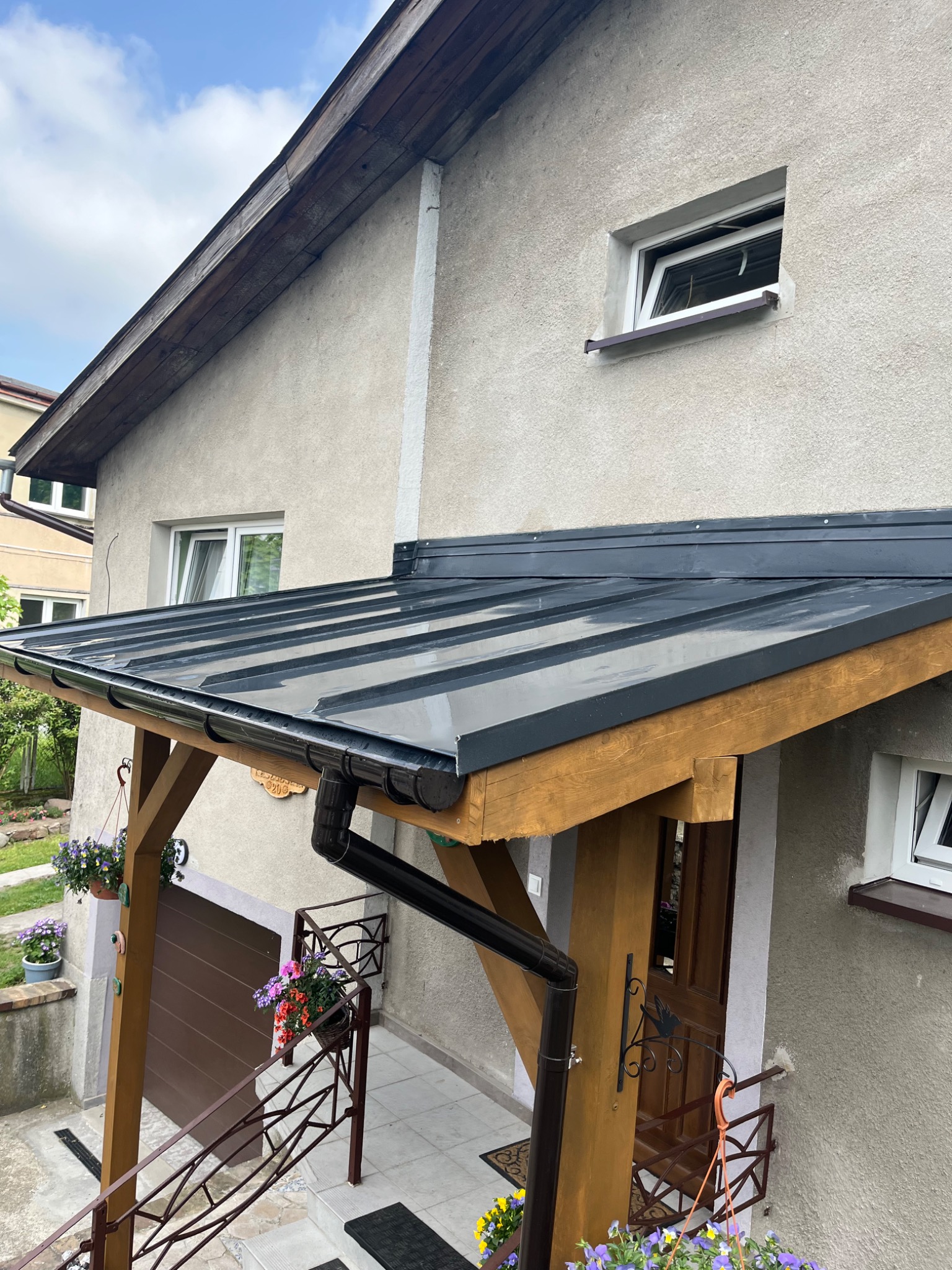 Zadaszenie nad wejściem do domu z ciemnej blachy na drewnianej konstrukcji, rynna spustowa z PCV, widok z dołu.