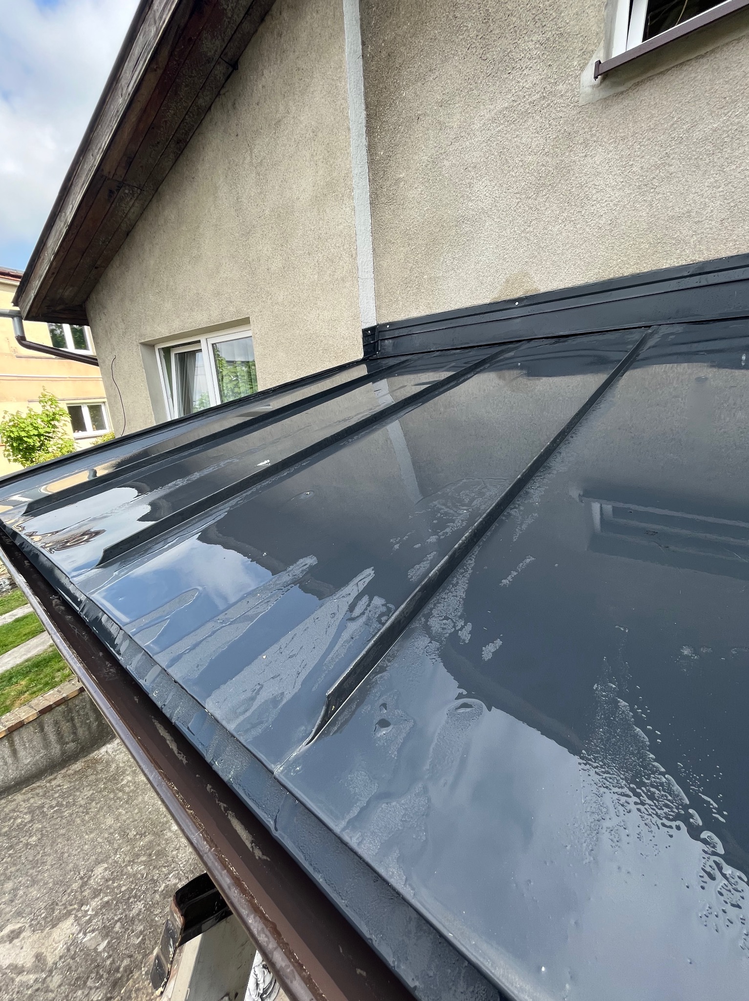 Błyszczący, nowy, grafitowy dach panelowy z widocznymi kroplami wody po deszczu, brązowa rynna wzdłuż krawędzi dachu, jasna elewacja budynku w tle.