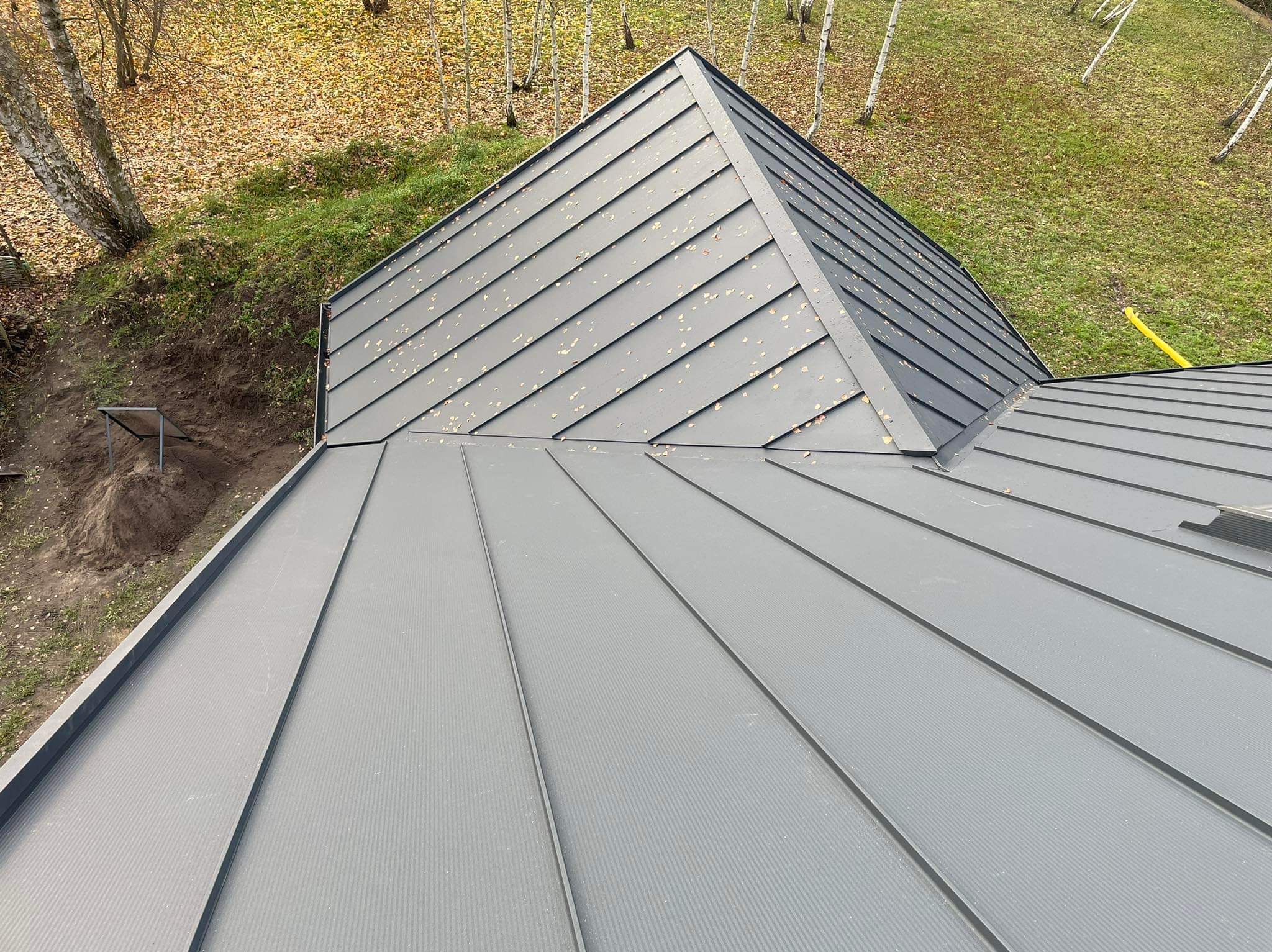 Szary, blaszany dach z widocznymi łączeniami blach na tle drzew i trawy, z perspektywy z góry.