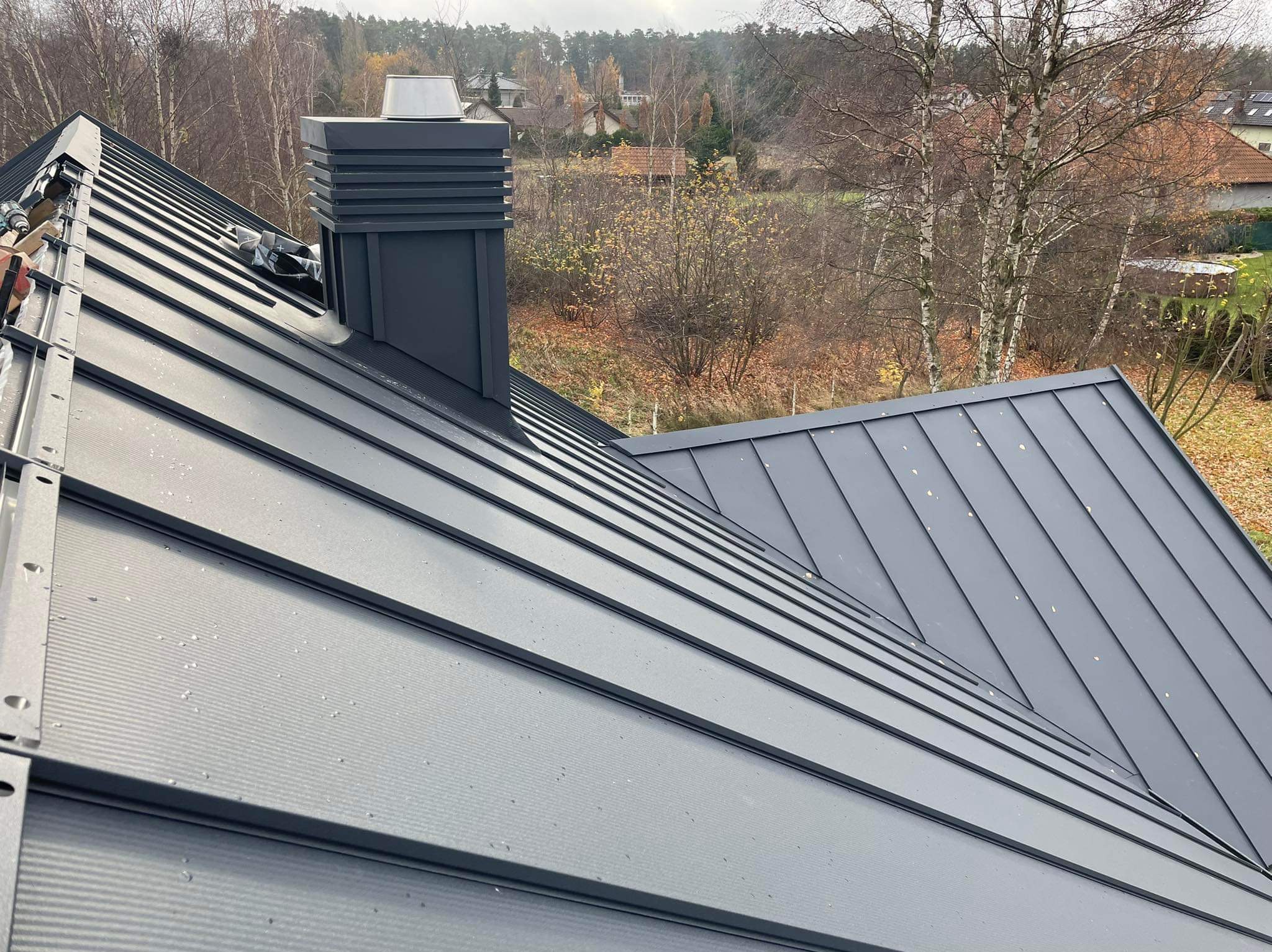 Nowoczesny, szary dach z blachy na rąbek stojący, z widocznym kominem i elementami montażowymi, na tle jesiennego krajobrazu.