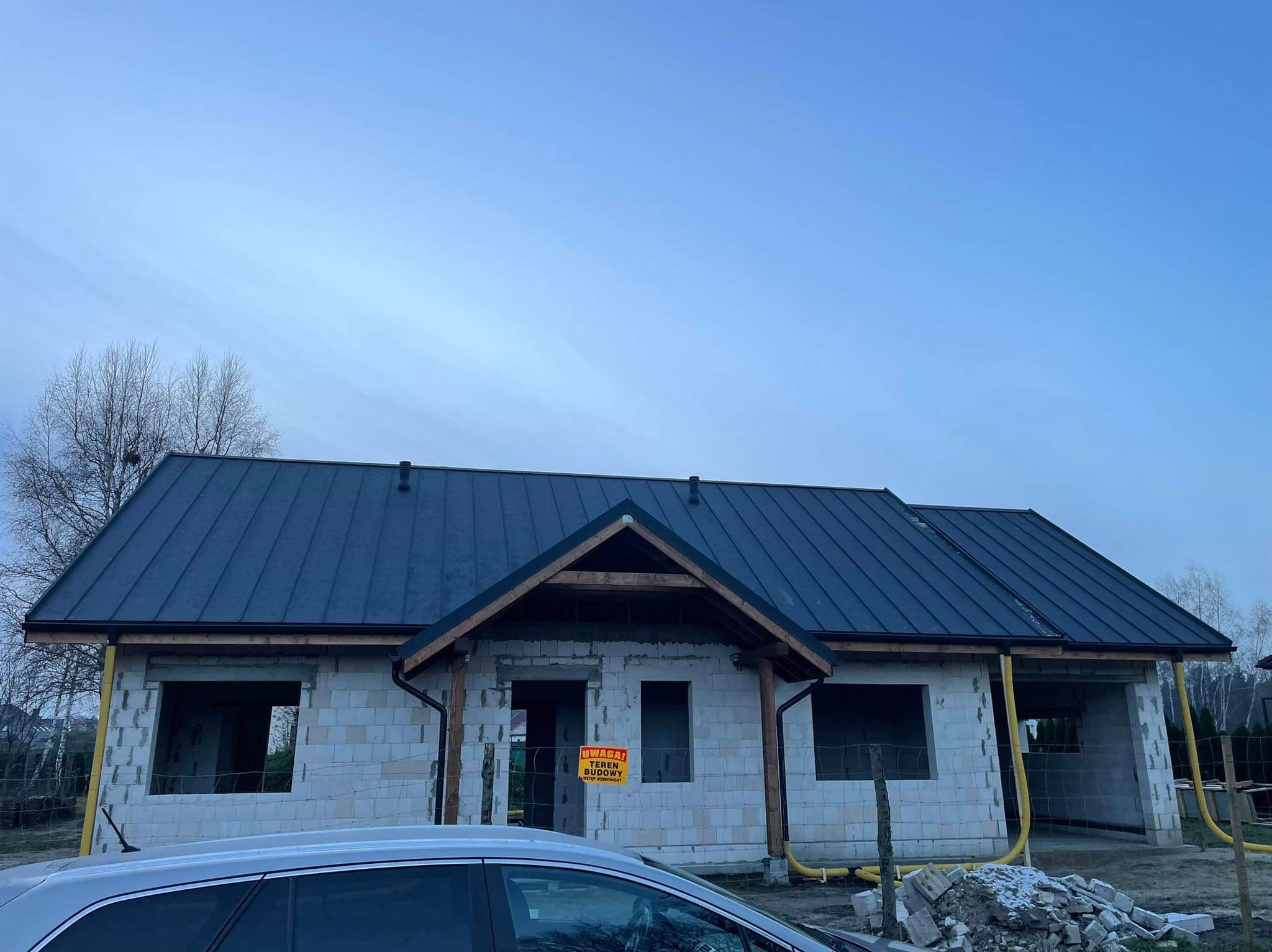 Niedokończony dom z ciemnym, metalowym dachem, widoczne rynny i obróbki blacharskie, żółte rury wentylacyjne na elewacji, tablica ostrzegawcza 'Teren budowy'.