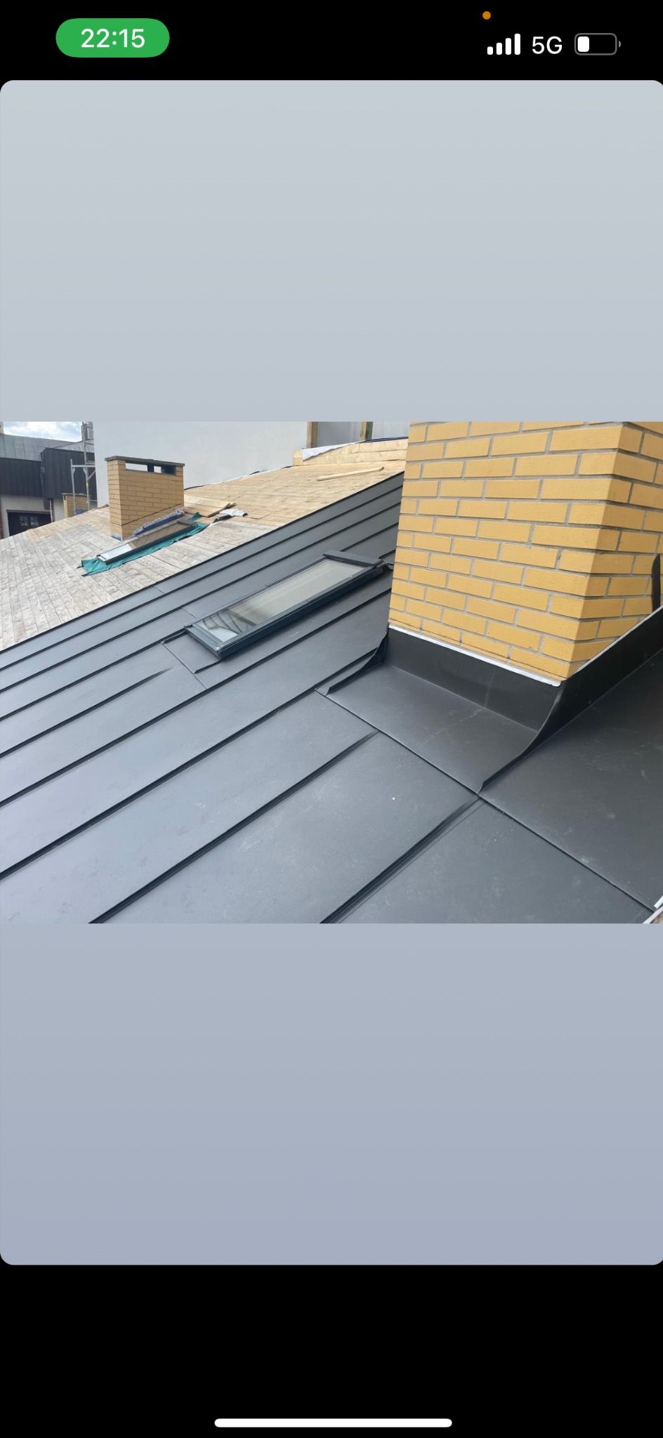 Czarny dach z blachy na rąbek stojący z oknem dachowym i ceglanym kominem, w tle inny dach w trakcie budowy.