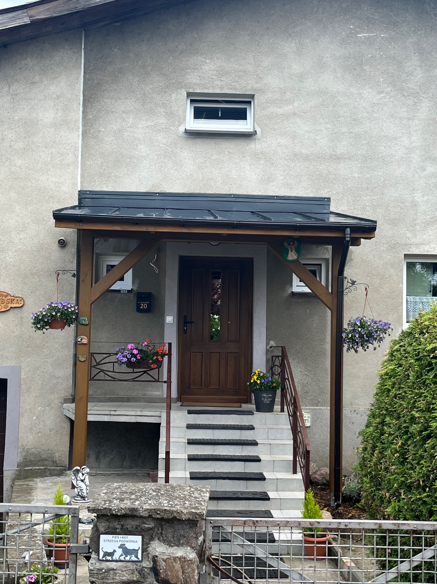Zadaszone wejście do domu z drewnianą konstrukcją, czarną blachodachówką, wiszącymi kwiatami i schodami z antypoślizgowymi nakładkami.