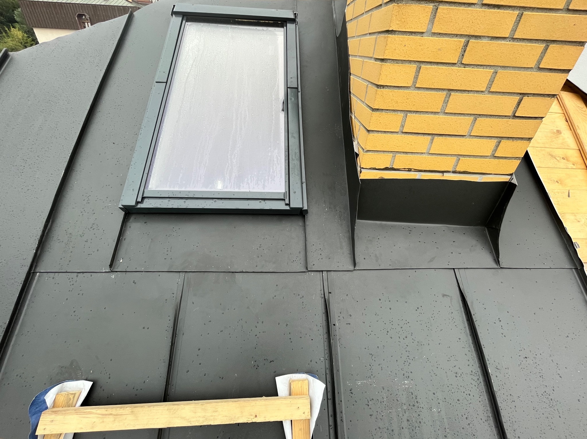 Nowoczesne, grafitowe pokrycie dachowe z blachy na rąbek stojący, z oknem dachowym i kominem z żółtej cegły, widoczne krople deszczu.