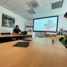 BE- Dach (VELOS) Emil Szukiełowicz - Prezentacja IKO Premium dakbanen z wizualizacją warstw dachowych na ekranie projektora w sali konferencyjnej, widoczny mężczyzna z laptopem i notatnik z zapiskami.