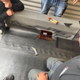 BE- Dach (VELOS) Emil Szukiełowicz - Przygotowanie papy termozgrzewalnej na stole warsztatowym, widoczne narzędzia: nóż dekarski, dociskacz i szablon, trzech dekarzy w trakcie pracy.