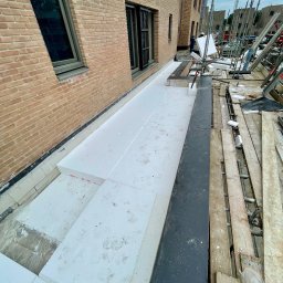 BE- Dach (VELOS) Emil Szukiełowicz - Białe płyty izolacyjne ułożone na dachu budynku w trakcie prac budowlanych, widoczne rusztowania i fragment elewacji z cegły.