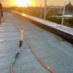 Praca dekarska na płaskim dachu podczas zachodu słońca: palnik dekarski w użyciu, widoczne zgrzewane pasy papy oraz rusztowanie na krawędzi dachu, w tle zabudowania miejskie.