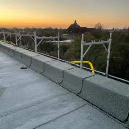 Nowy, płaski dach pokryty papą termozgrzewalną z obróbkami blacharskimi w kolorze szarym, widoczne rusztowanie na dachu i budynek w tle o zachodzie słońca.