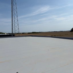 BE- Dach (VELOS) Emil Szukiełowicz - Jasnoszary, płaski dach budynku z widocznymi łączeniami, na tle błękitnego nieba i krajobrazu z drzewem i wieżą telekomunikacyjną w oddali. Widok z góry.