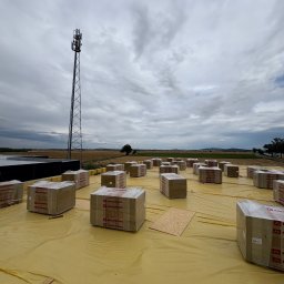 BE- Dach (VELOS) Emil Szukiełowicz - Paczki wełny mineralnej Rockwool na żółtej membranie dachowej, przygotowane do ocieplenia dachu. W tle wieża telekomunikacyjna i pochmurne niebo.