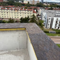 BE- Dach (VELOS) Emil Szukiełowicz - Fragment dachu z płyt OSB i ociepleniem, widok z góry na tle zabudowy miejskiej i zieleni. Widoczne detale łączeń i faktura materiałów.