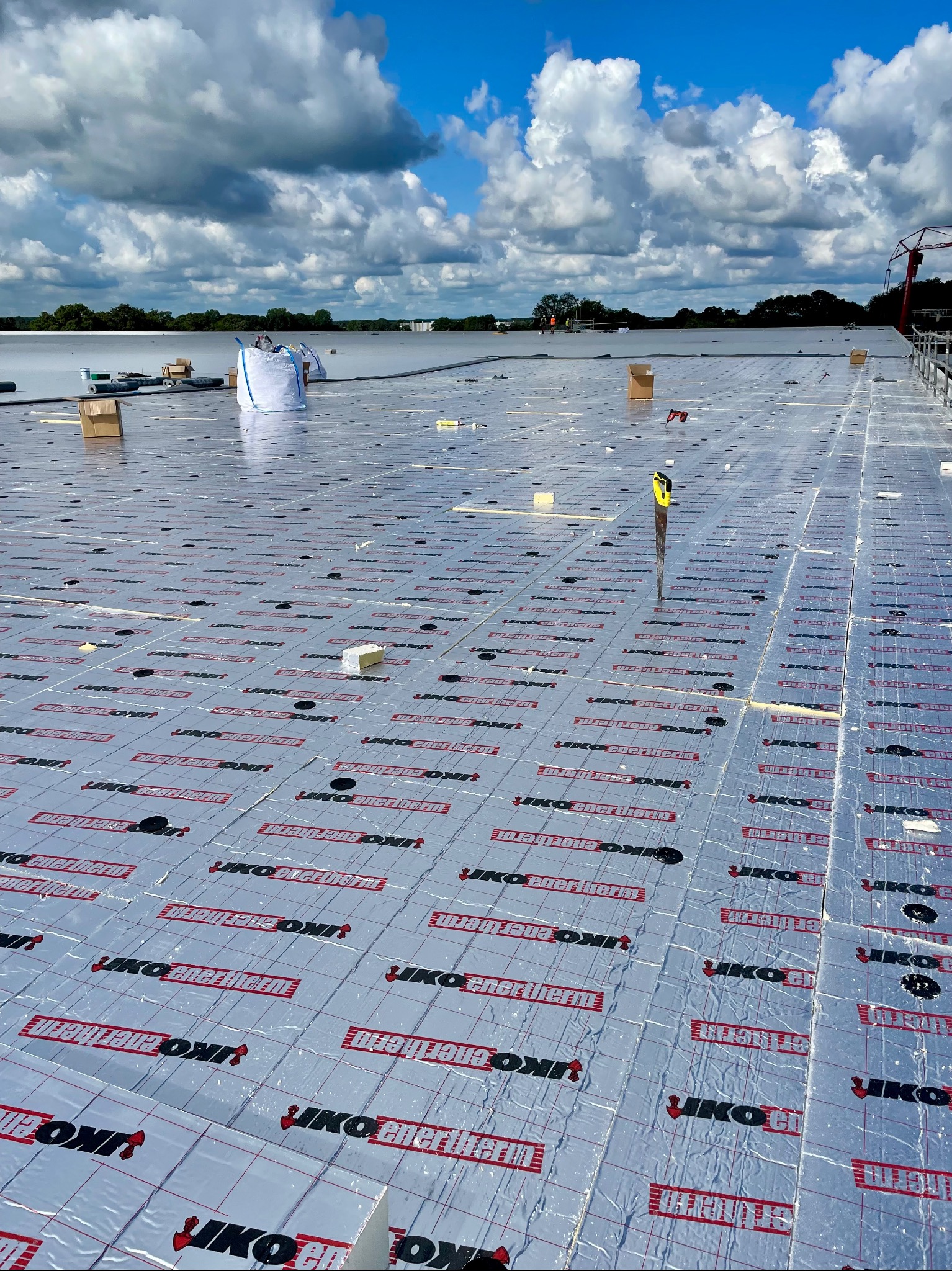 Płyty izolacyjne IKO enertherm układane na dachu, widoczny nóż do cięcia izolacji, w tle błękitne niebo z chmurami i fragment dźwigu.