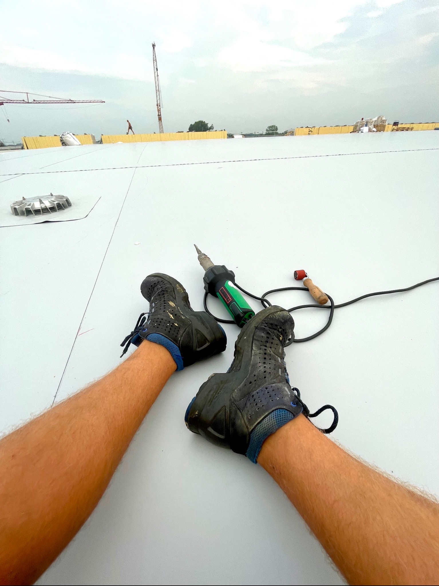 Przerwa w pracy na dachu: Widok z perspektywy pracownika na płaskiej powierzchni dachu, obok leżą buty robocze, zgrzewarka do papy i wałek dociskowy, w tle dźwigi budowlane i pochmurne niebo.