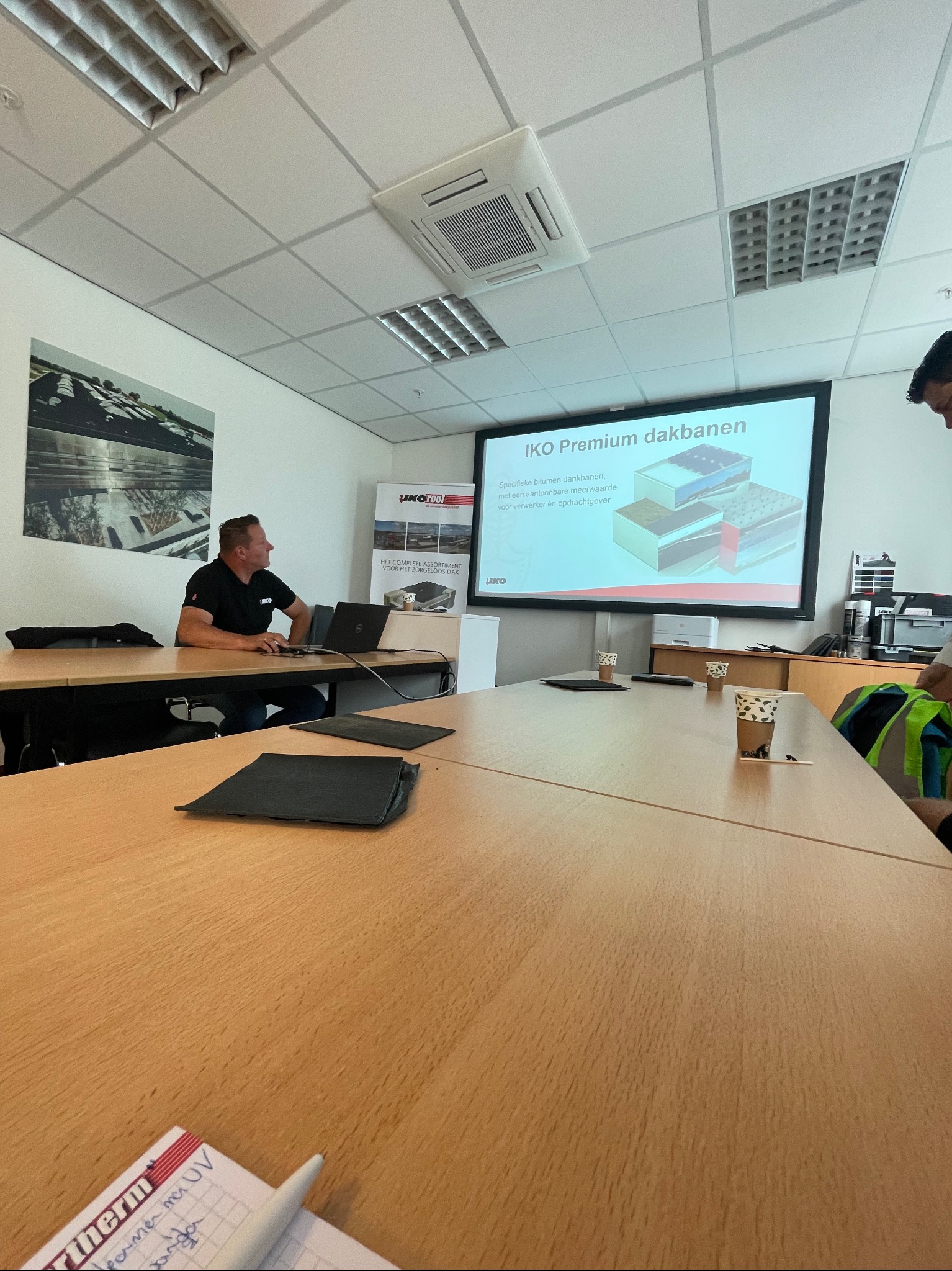 Prezentacja IKO Premium dakbanen z wizualizacją warstw dachowych na ekranie projektora w sali konferencyjnej, widoczny mężczyzna z laptopem i notatnik z zapiskami.