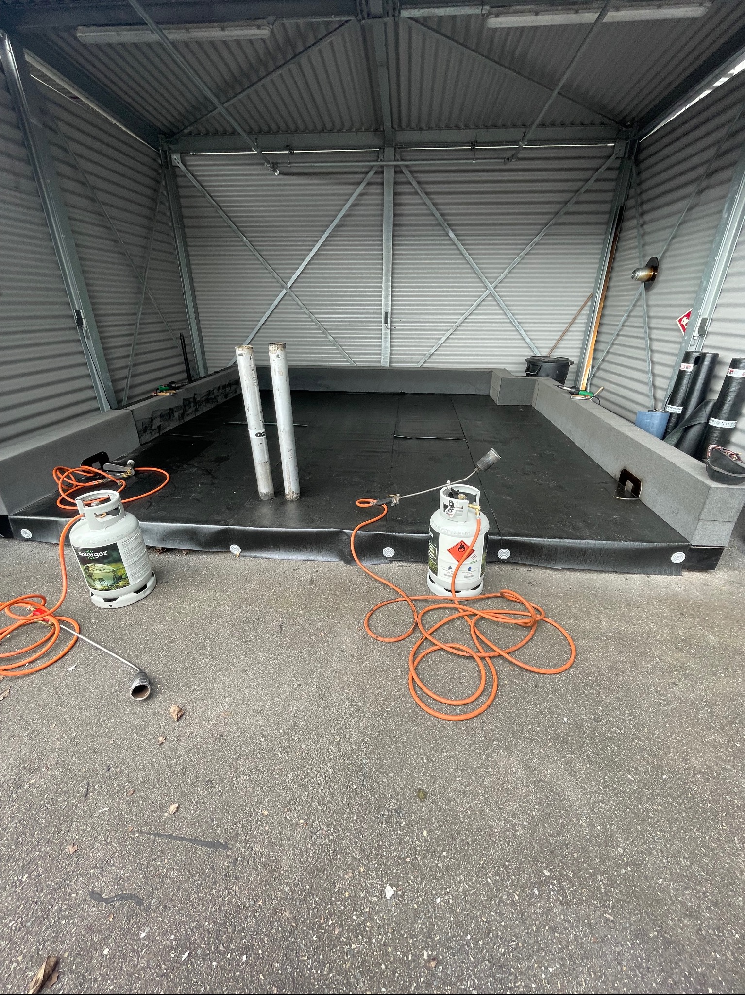 Montaż izolacji bitumicznej na dachu garażu blaszanego, widoczne butle z gazem i palnik dekarski.