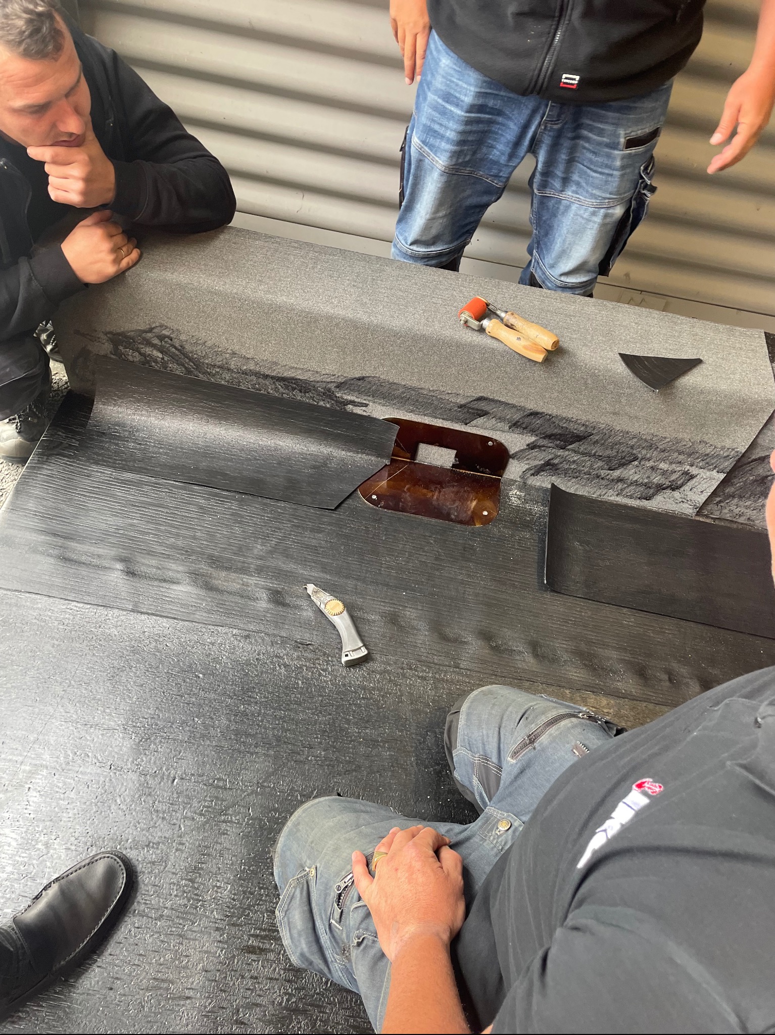 Przygotowanie papy termozgrzewalnej na stole warsztatowym, widoczne narzędzia: nóż dekarski, dociskacz i szablon, trzech dekarzy w trakcie pracy.