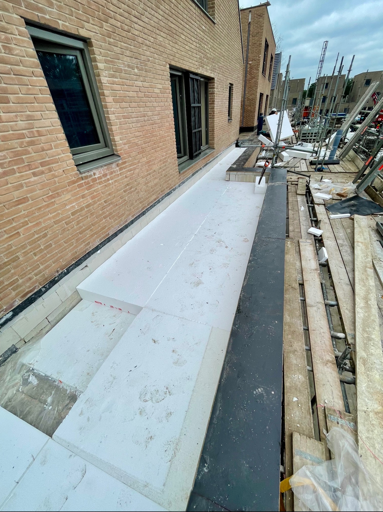 Białe płyty izolacyjne ułożone na dachu budynku w trakcie prac budowlanych, widoczne rusztowania i fragment elewacji z cegły.