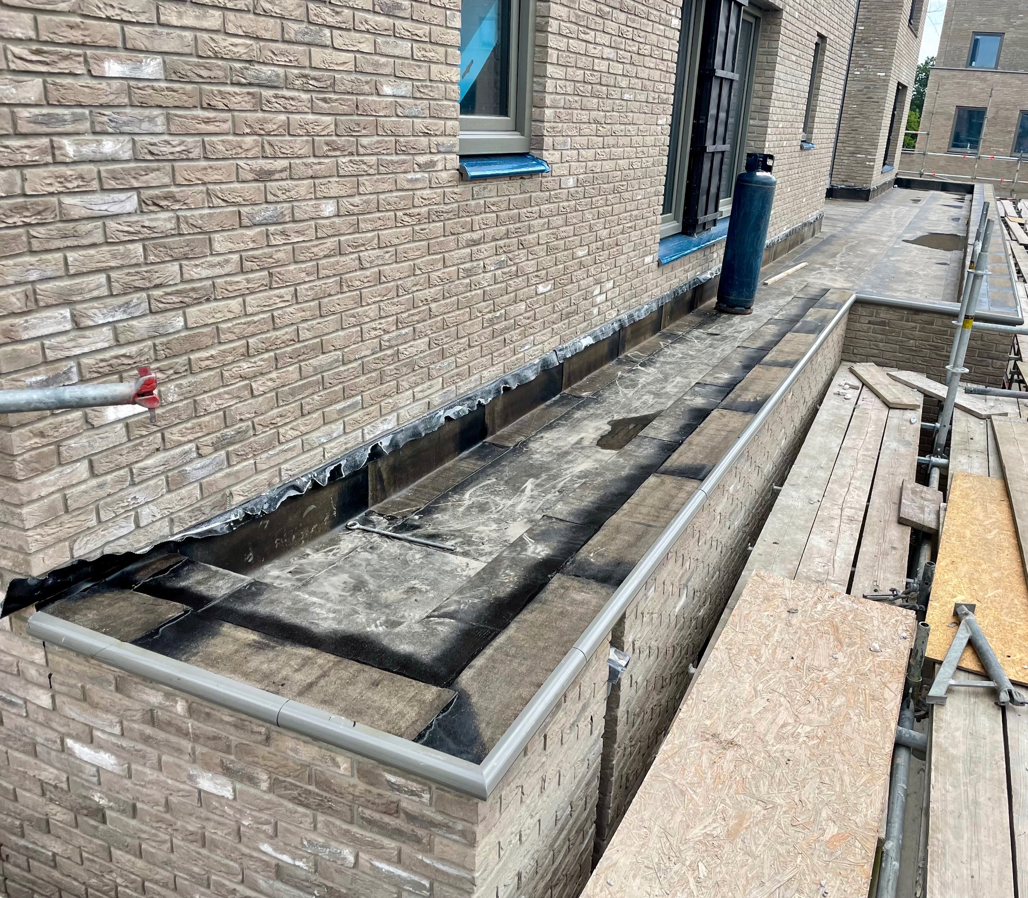Balkon wykończony cegłą klinkierową z hydroizolacją, widoczne rusztowania i butla gazowa.