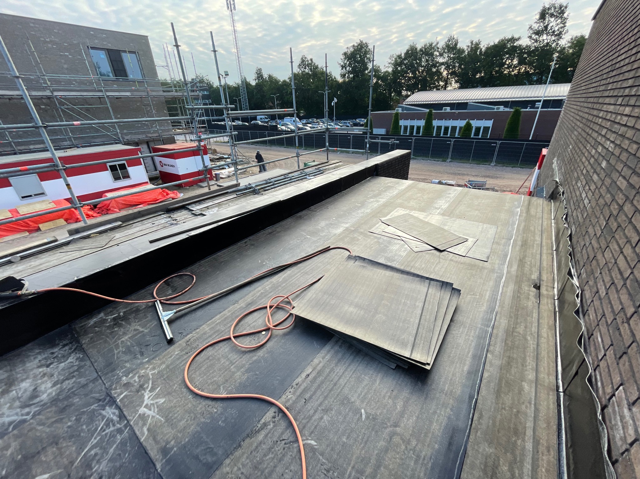 Fragment dachu pokrytego papą termozgrzewalną w trakcie prac dekarskich, widoczne narzędzia, bryły papy oraz budynek w tle z rusztowaniami.