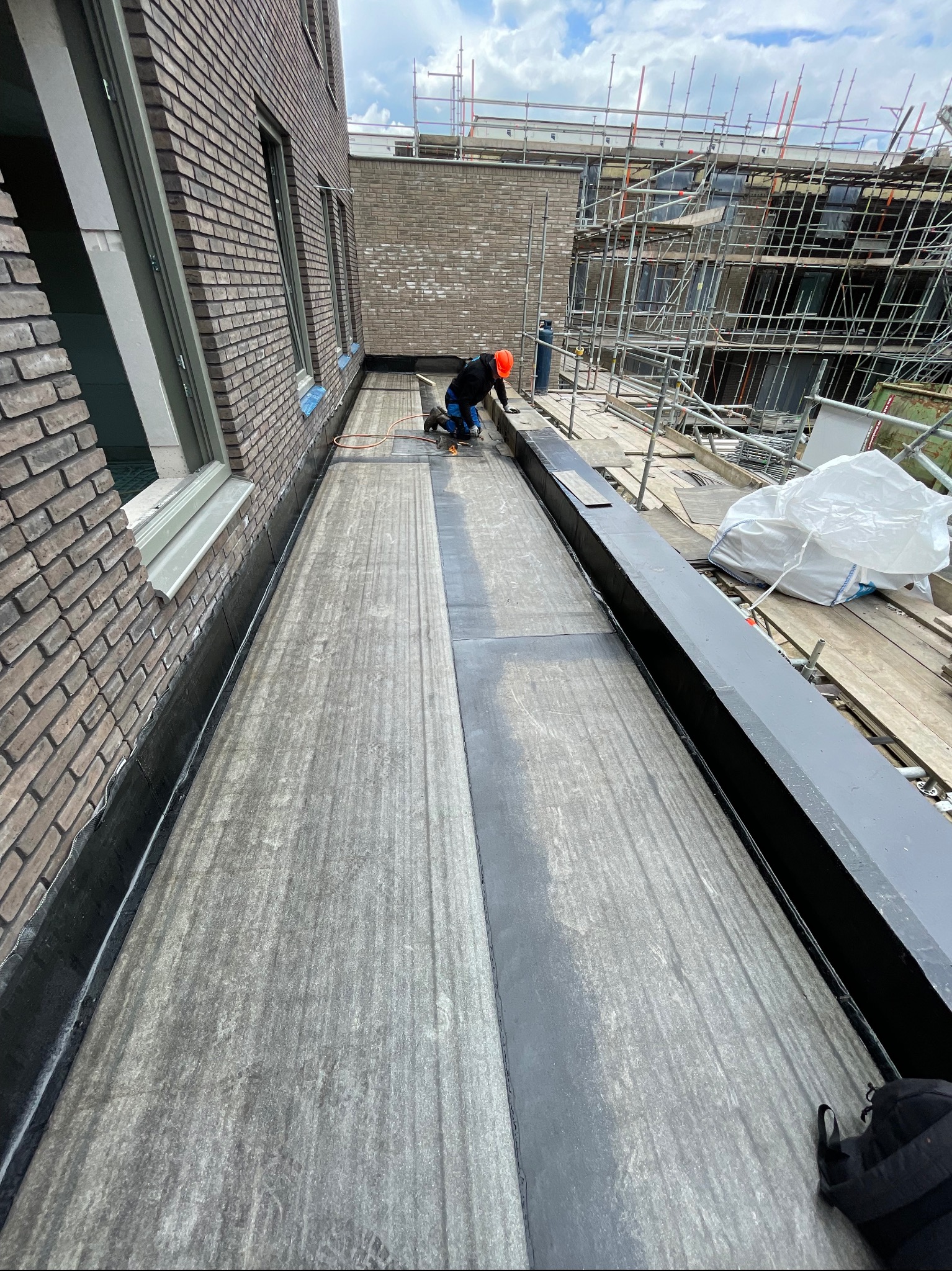 Praca na dachu budynku z cegły: dekarz w pomarańczowym kasku uszczelnia papę termozgrzewalną za pomocą palnika, widoczne rusztowania i materiały budowlane w tle.