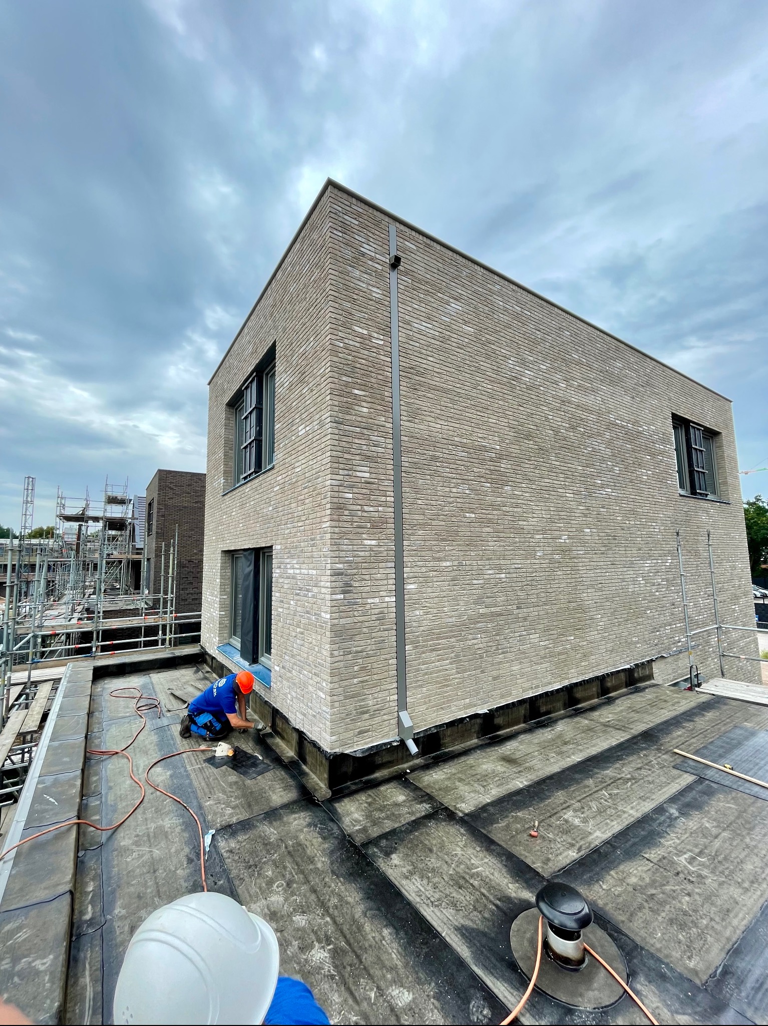 Pracownik w pomarańczowym kasku, w trakcie uszczelniania dachu z papy na nowym budynku z jasnej cegły, widoczne narzędzia i elementy instalacji wentylacyjnej, w tle rusztowania i pochmurne niebo.