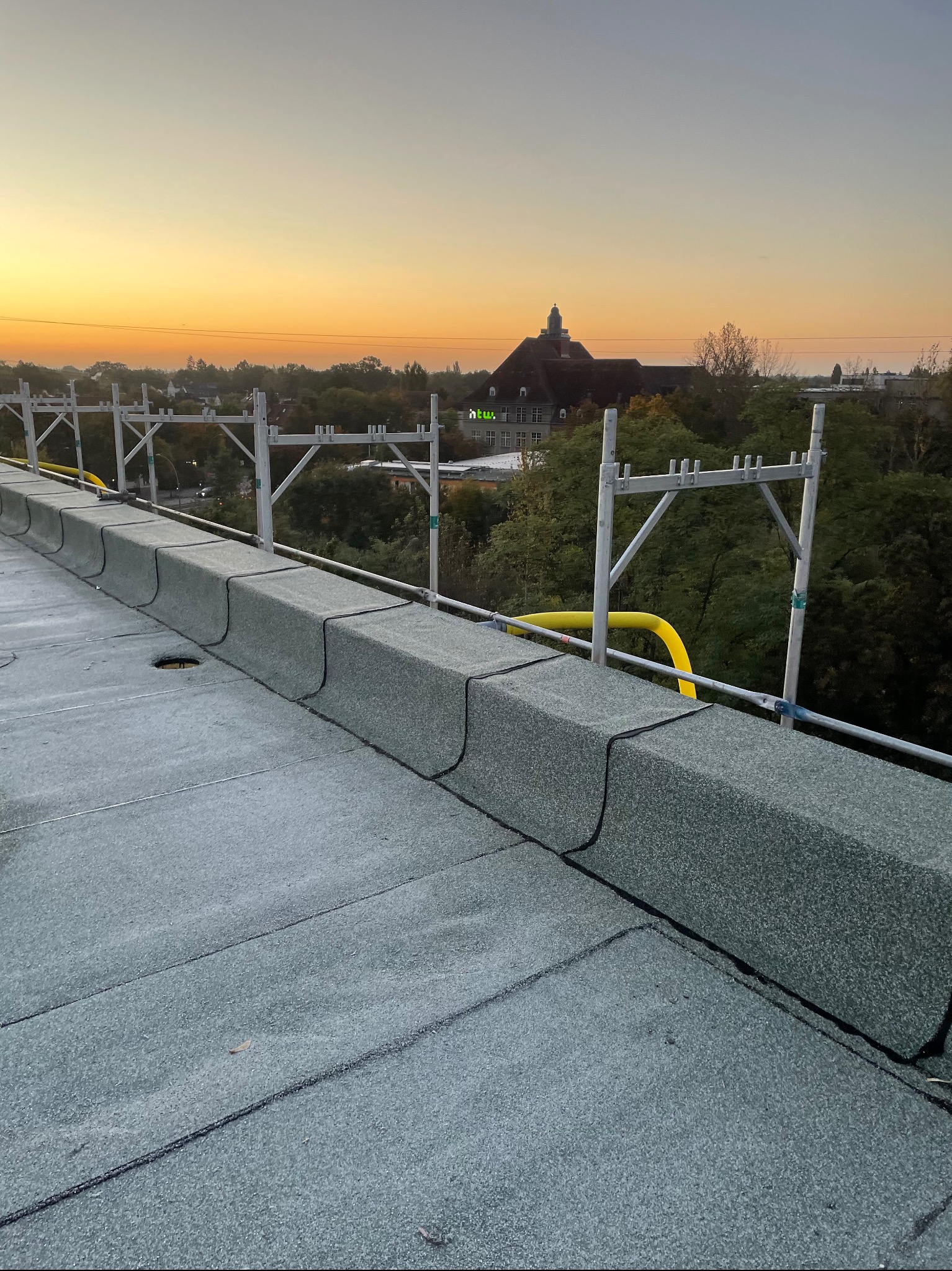 Nowy, płaski dach pokryty papą termozgrzewalną z obróbkami blacharskimi w kolorze szarym, widoczne rusztowanie na dachu i budynek w tle o zachodzie słońca.