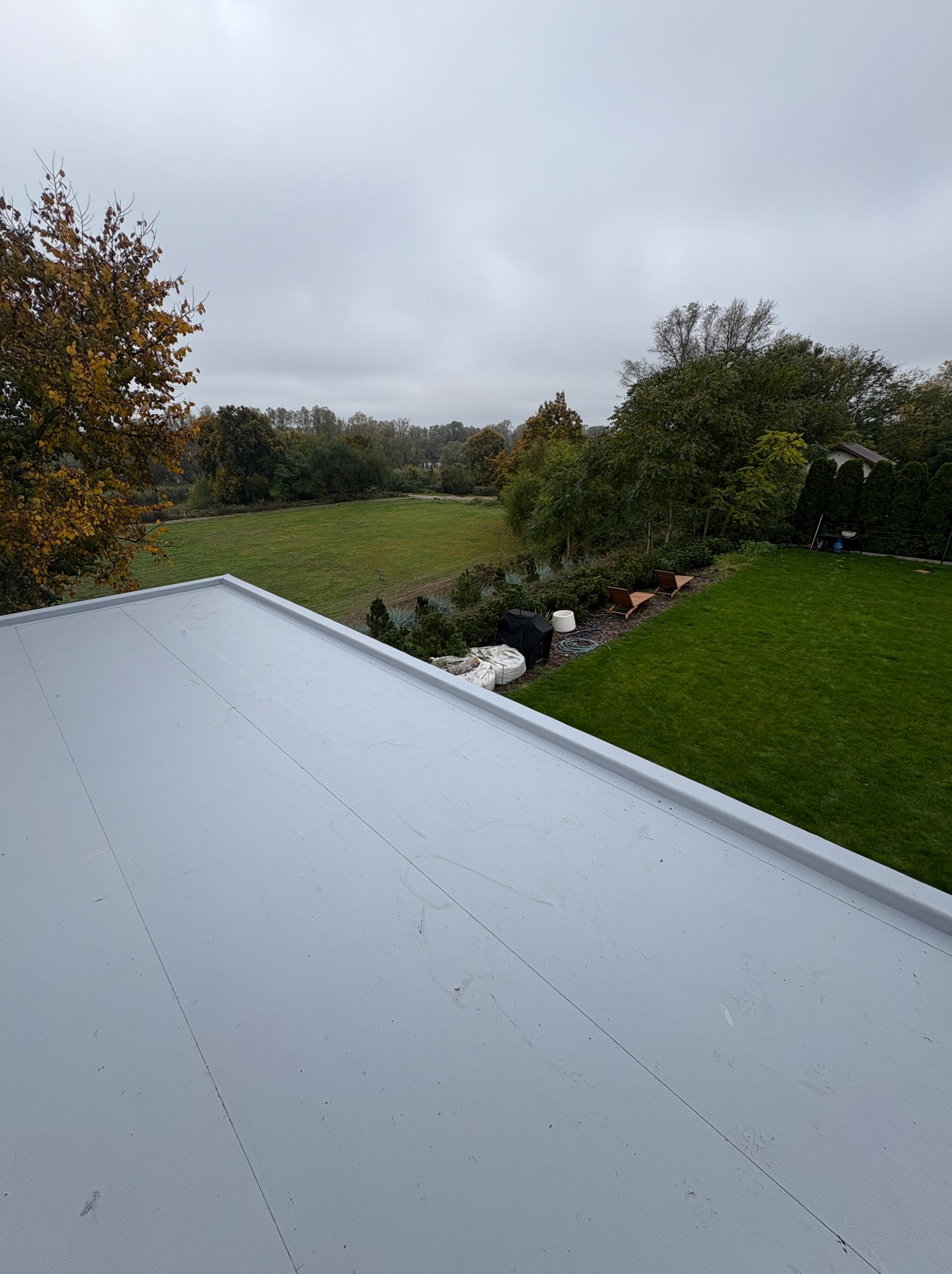 Szary, płaski dach z widokiem na zielony ogród i drzewa w tle, sfotografowany w pochmurny dzień. Widoczne łączenia papy na dachu.