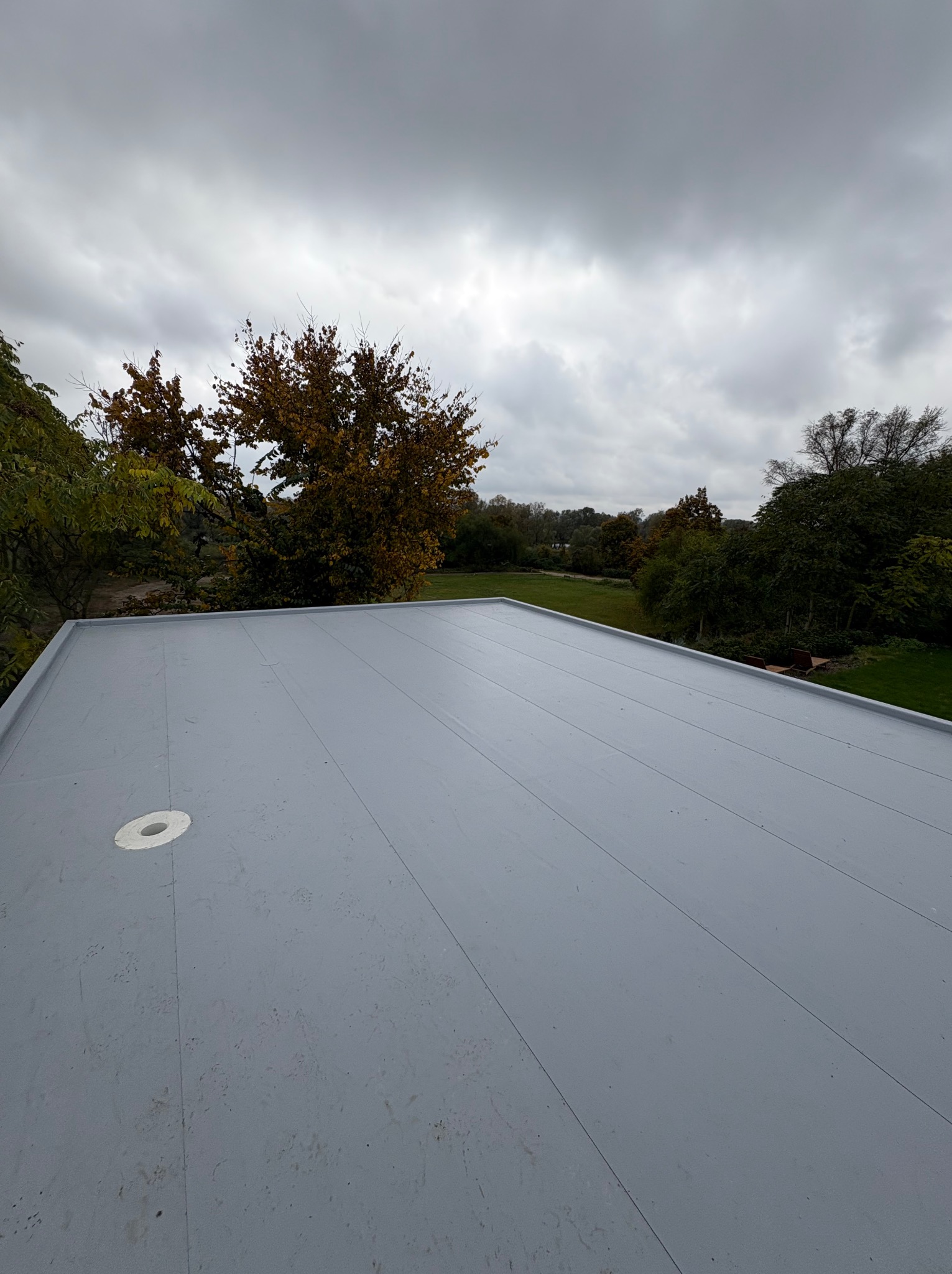 Szary, płaski dach z widocznym odpływem wody, na tle jesiennego krajobrazu i pochmurnego nieba. Minimalistyczny design, równa powierzchnia, brak widocznych łączeń.