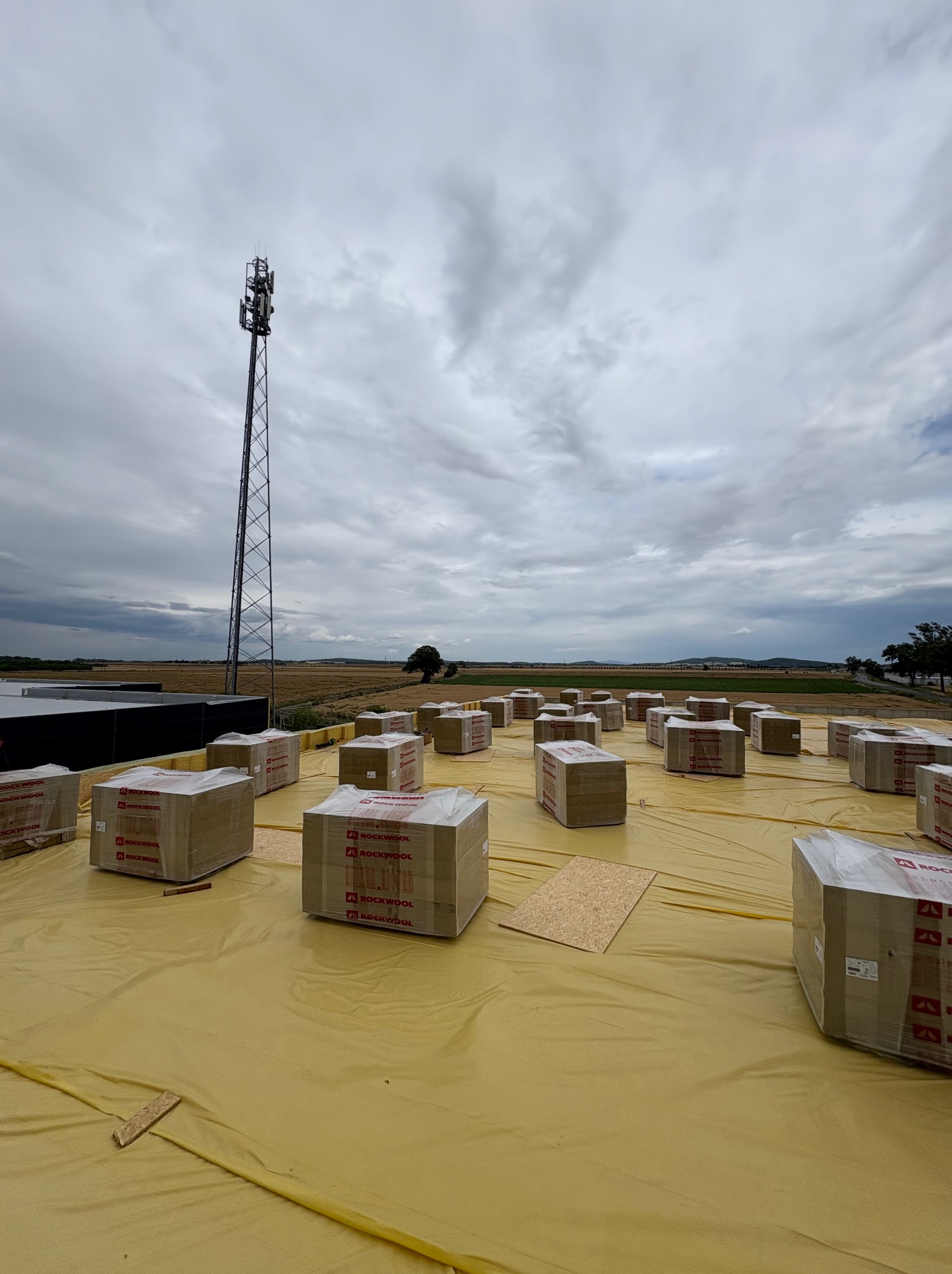 Paczki wełny mineralnej Rockwool na żółtej membranie dachowej, przygotowane do ocieplenia dachu. W tle wieża telekomunikacyjna i pochmurne niebo.