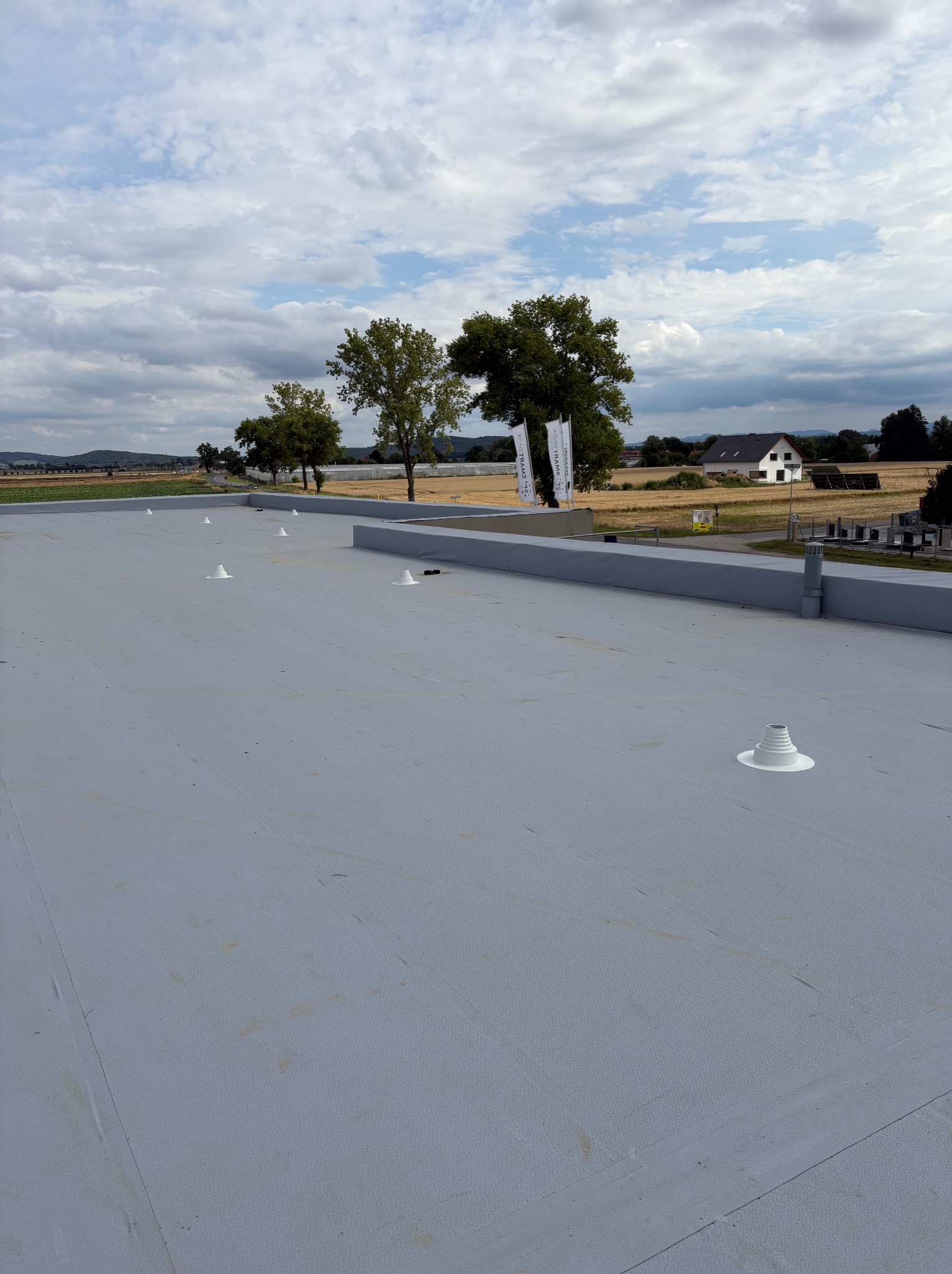 Szary, płaski dach z elementami wentylacyjnymi na tle krajobrazu z drzewami i budynkiem. Widok z góry, oświetlenie dzienne, lekko pochmurne niebo.