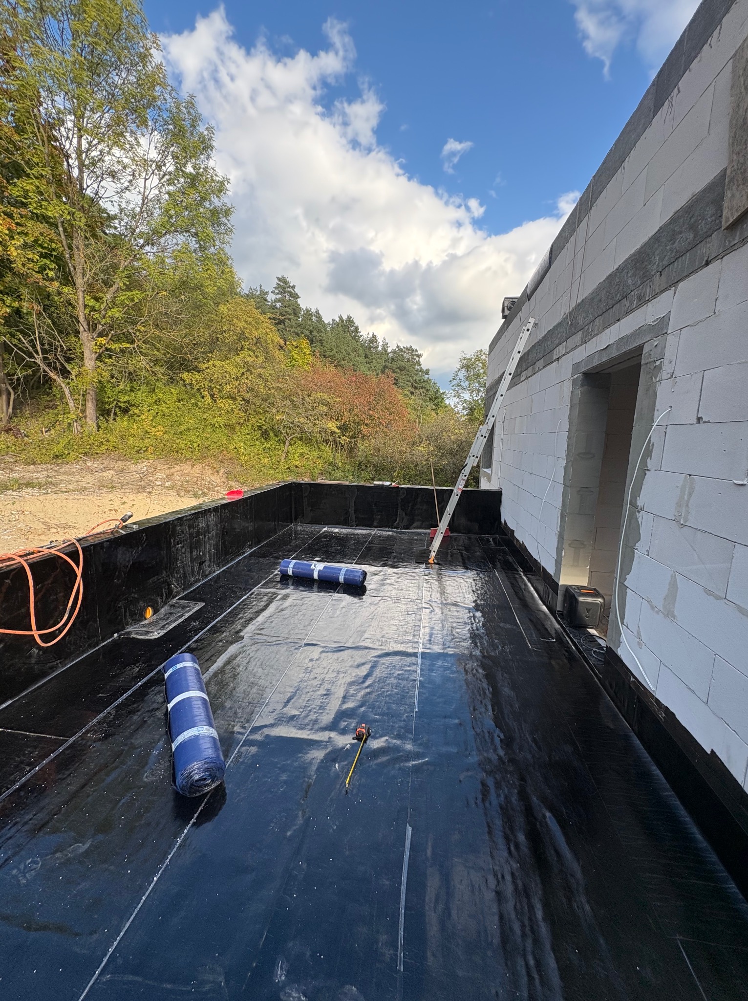 Czarna hydroizolacja dachu płaskiego w trakcie montażu, widoczne rolki materiału, drabina oparta o ścianę budynku z betonu komórkowego, błękitne niebo.