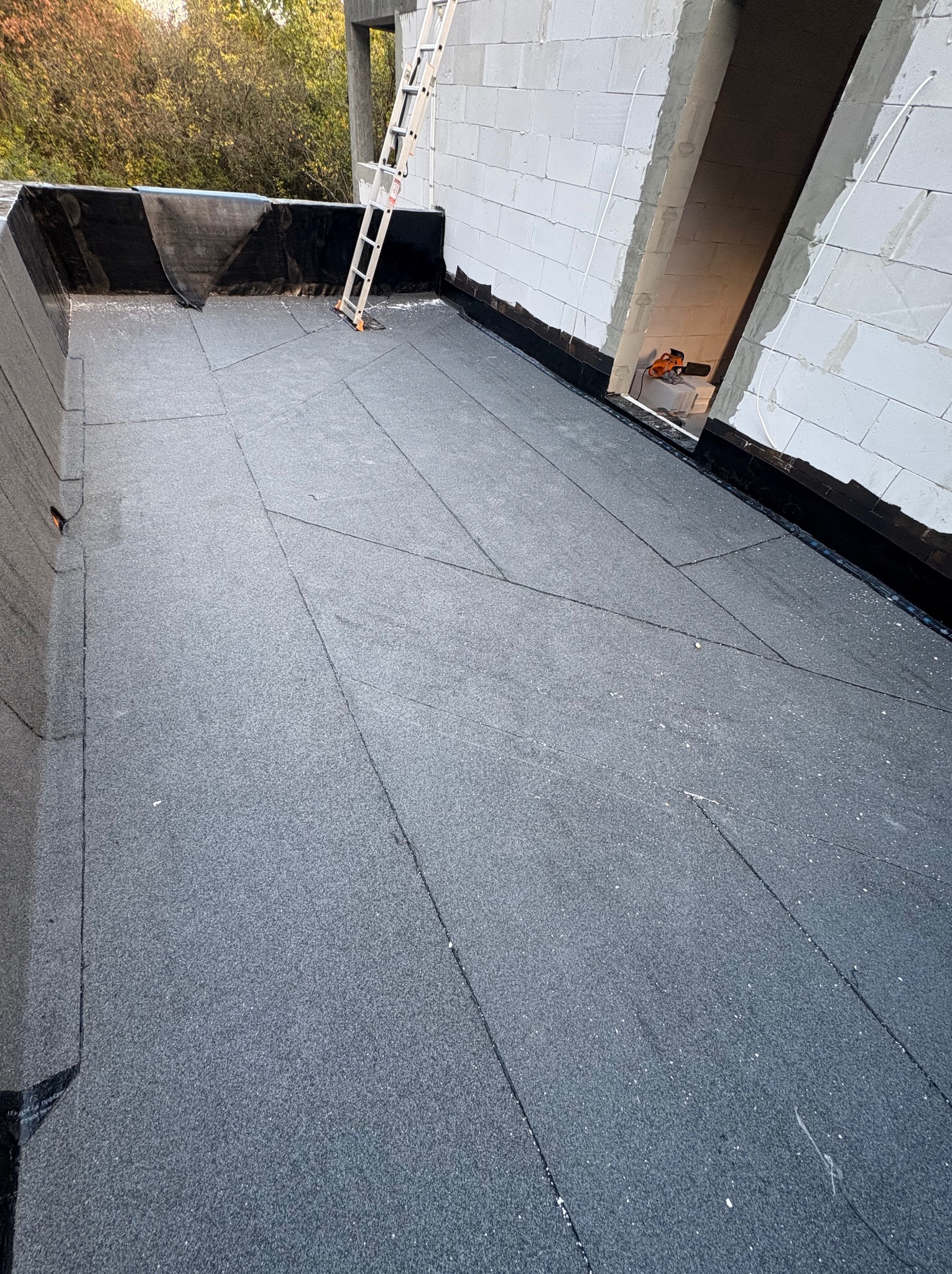 Szary, płaski dach pokryty papą termozgrzewalną z widoczną drabiną i fragmentem ściany budynku w trakcie budowy. Widoczne łączenia papy.