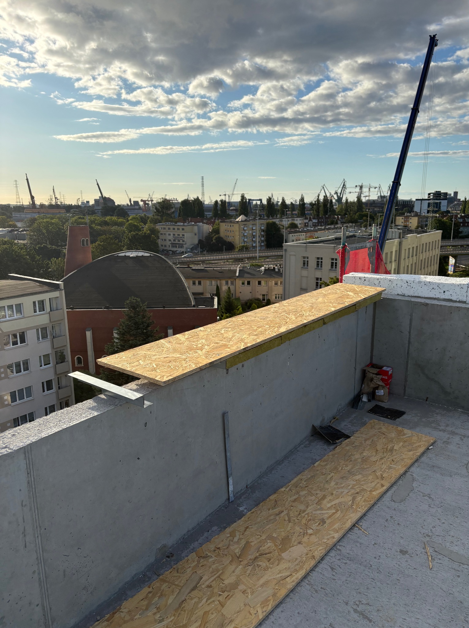 Montaż płyty OSB na betonowej ścianie budynku w trakcie budowy. W tle widok na miasto z dźwigami i budynkami. Częściowo widoczne niebo z chmurami.