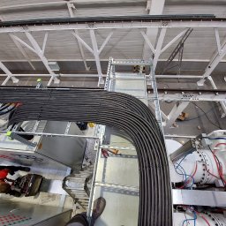 EL-DAM - Widok z góry na trasę kablową z ułożonymi kablami w hali przemysłowej, widoczne elementy konstrukcji dachu i fragment maszyny.