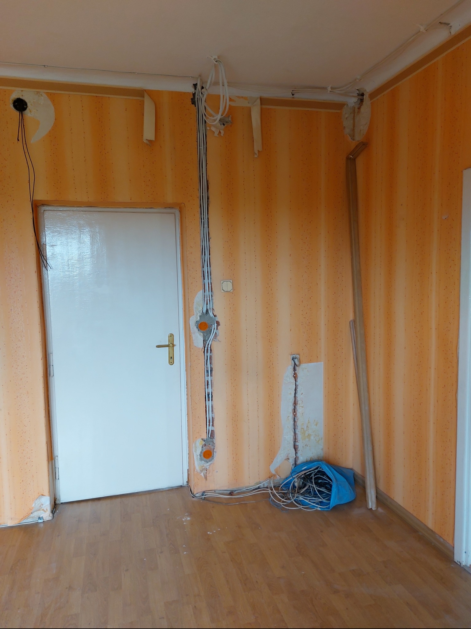 Pokój w trakcie remontu z widocznymi przewodami elektrycznymi wychodzącymi ze ściany, puszkami instalacyjnymi i otworami po gniazdkach, na podłodze leżą zwinięte kable w niebieskiej torbie.