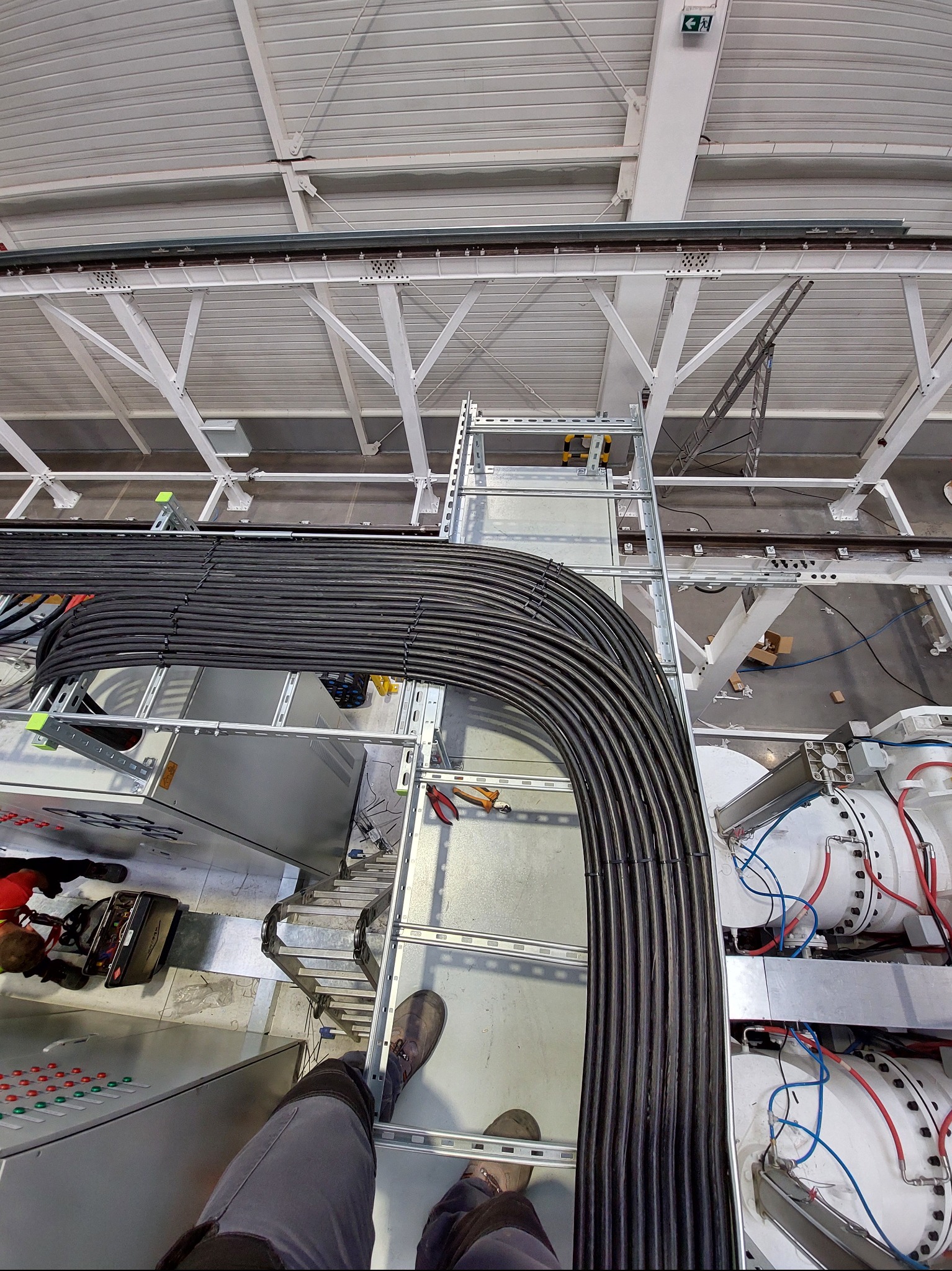 Widok z góry na trasę kablową z ułożonymi kablami w hali przemysłowej, widoczne elementy konstrukcji dachu i fragment maszyny.