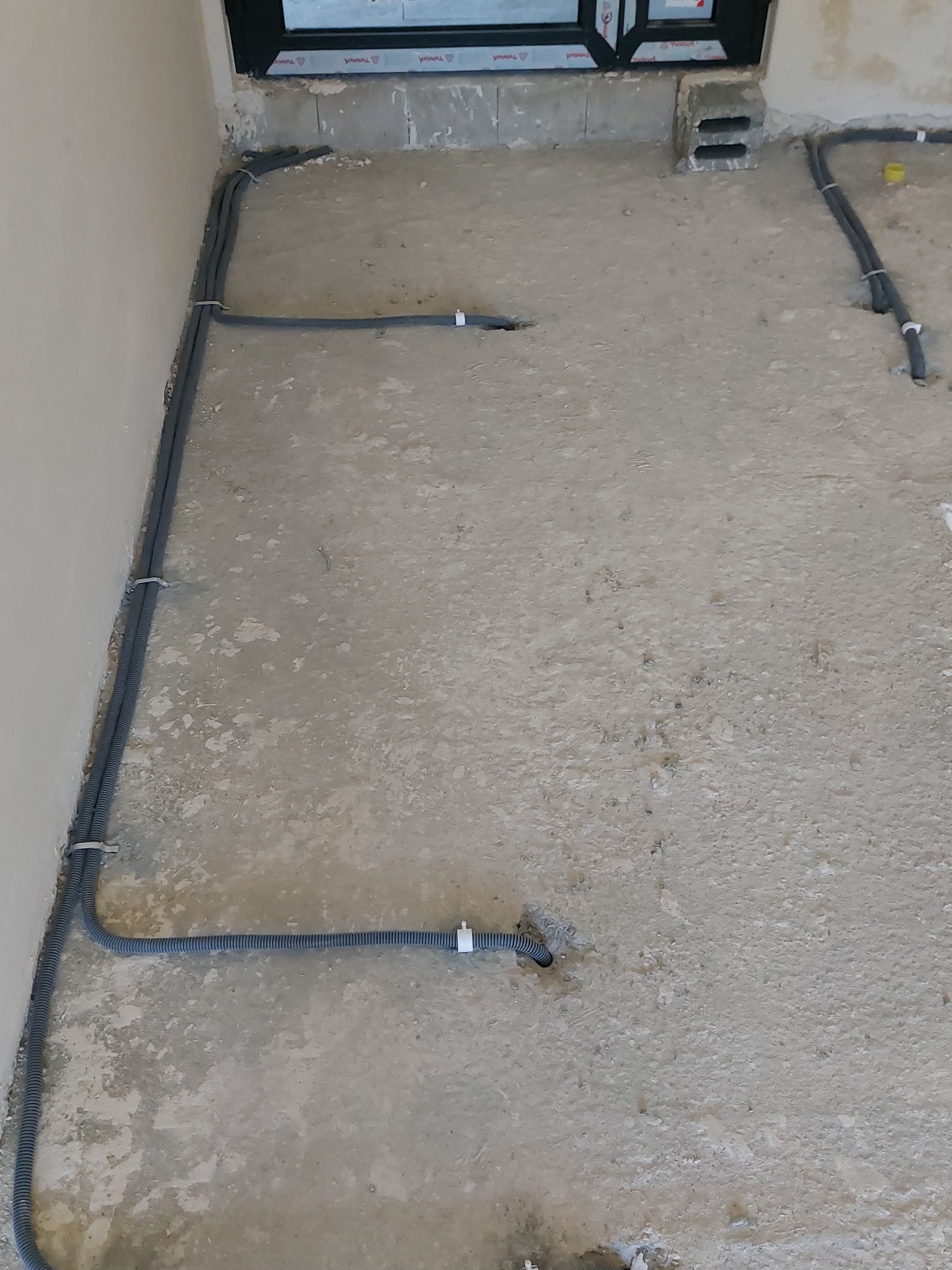 Szare rury karbowane prowadzone po szarej, betonowej podłodze w surowym stanie, przytwierdzone do podłoża białymi uchwytami, widoczne otwory w podłodze, okno w tle.