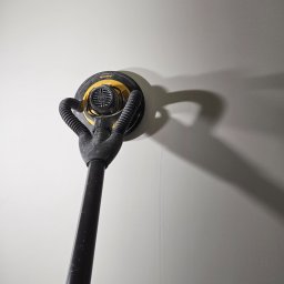 Daniel Wiński - Szlifierka oscylacyjna z odkurzaczem, marki Mirka, przyłożona do jasnej, gładkiej ściany. Widoczny cień urządzenia na ścianie. Na ścianie widoczne delikatne ślady po szlifowaniu.