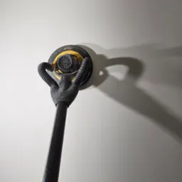 Szlifierka oscylacyjna z odkurzaczem, marki Mirka, przyłożona do jasnej, gładkiej ściany. Widoczny cień urządzenia na ścianie. Na ścianie widoczne delikatne ślady po szlifowaniu.