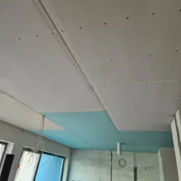 Montaż sufitu podwieszanego z płyt kartonowo-gipsowych na metalowej konstrukcji, widoczne szpachlowane łączenia płyt i instalacja elektryczna w surowym wnętrzu.