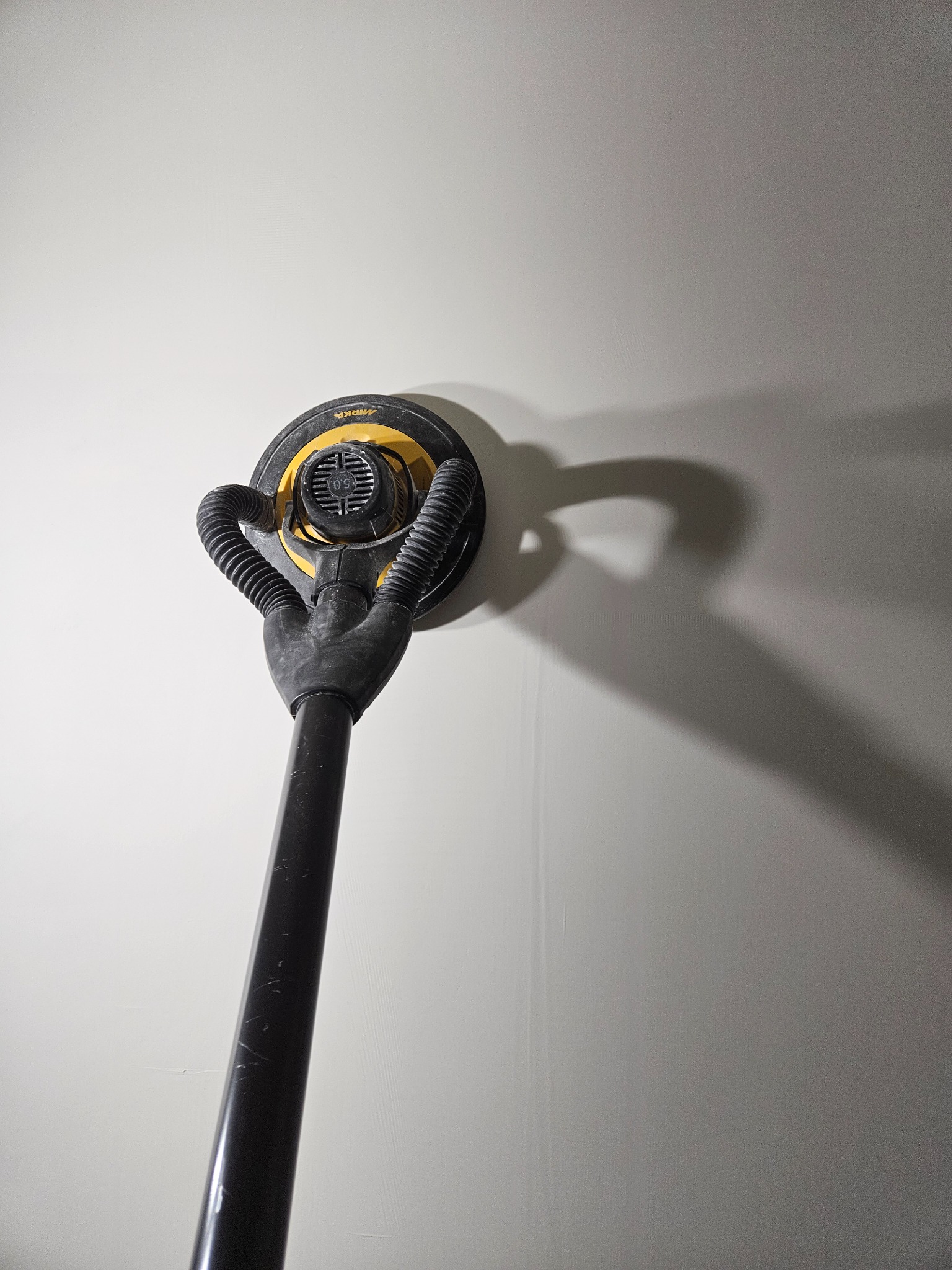 Szlifierka oscylacyjna z odkurzaczem, marki Mirka, przyłożona do jasnej, gładkiej ściany. Widoczny cień urządzenia na ścianie. Na ścianie widoczne delikatne ślady po szlifowaniu.