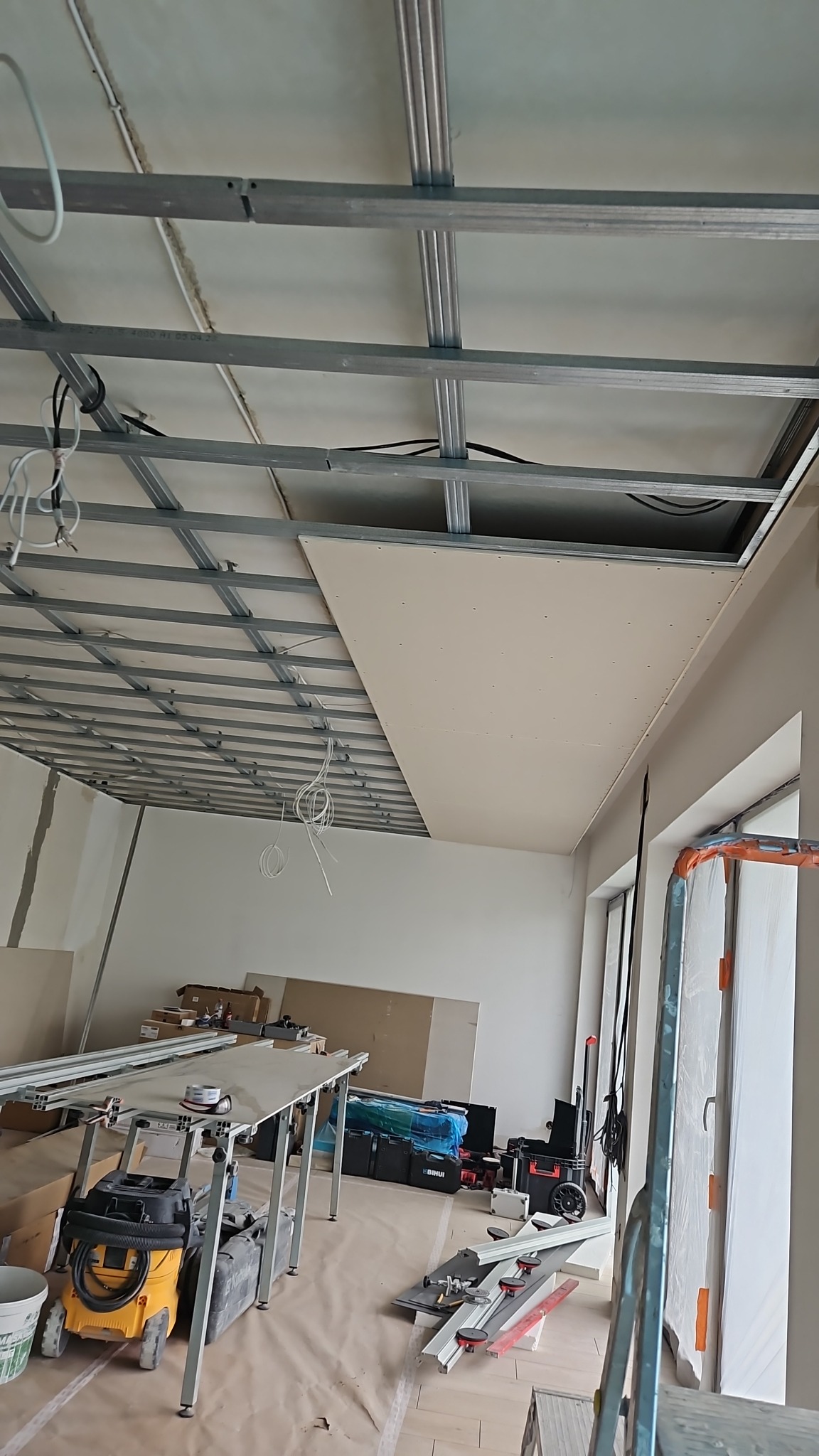 Montaż płyty karton gips na metalowym stelażu sufitu podwieszanego, widoczne narzędzia i materiały budowlane w pomieszczeniu w trakcie prac.
