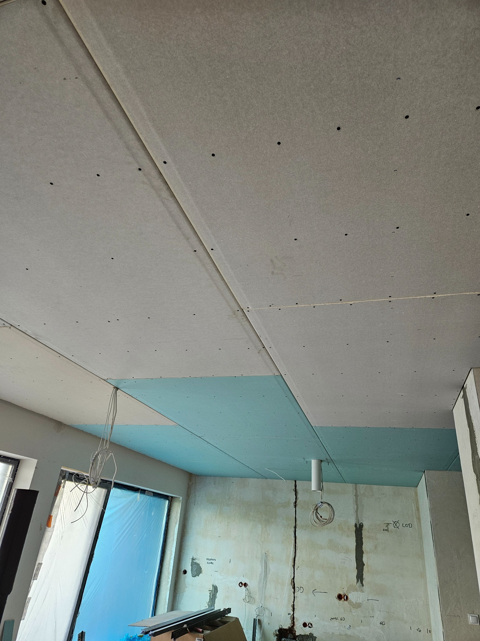 Montaż sufitu podwieszanego z płyt kartonowo-gipsowych na metalowej konstrukcji, widoczne szpachlowane łączenia płyt i instalacja elektryczna w surowym wnętrzu.